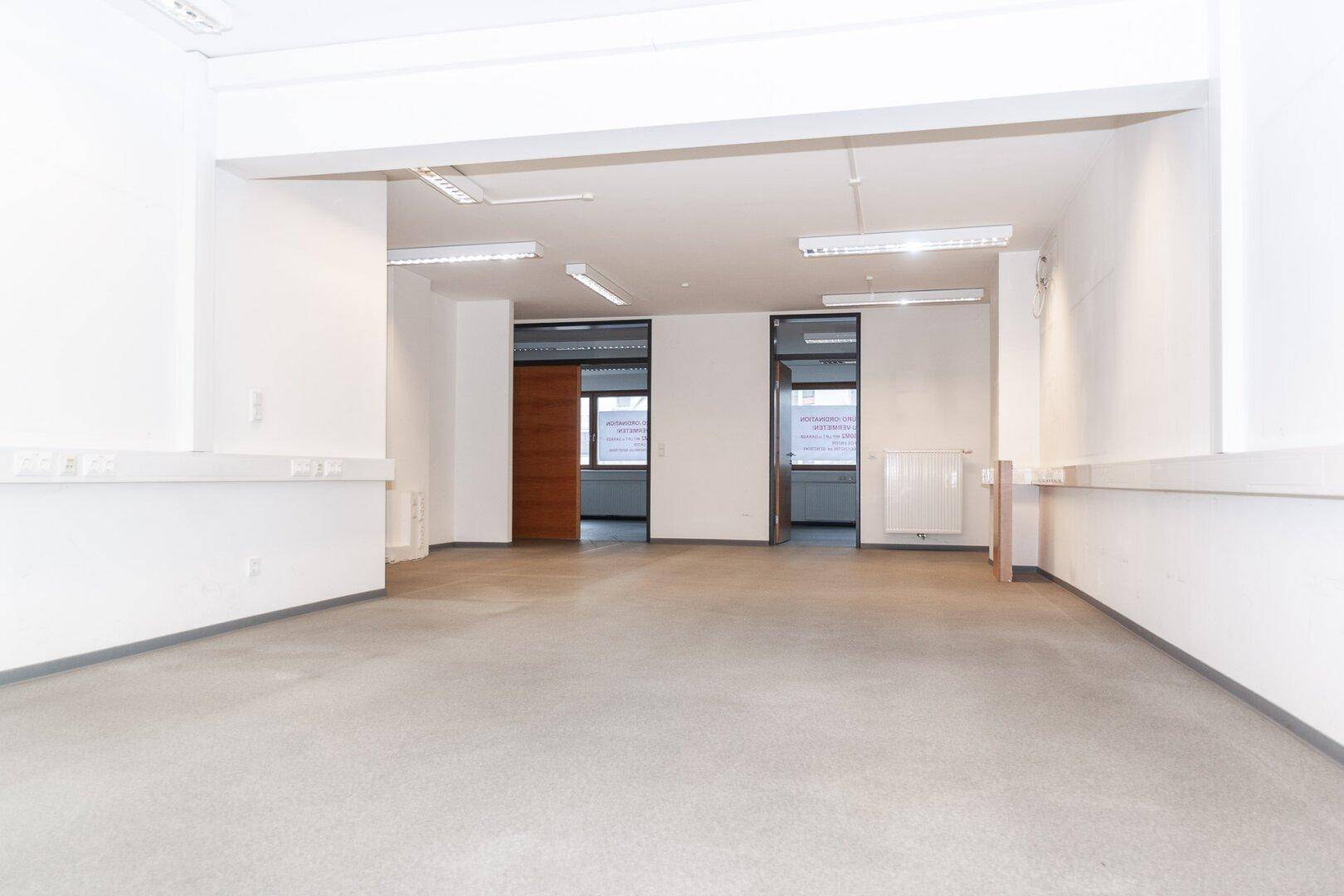 Weitläufiger, unmöblierter Bürobereich mit durchgehender Deckenbeleuchtung und mehreren Türen.