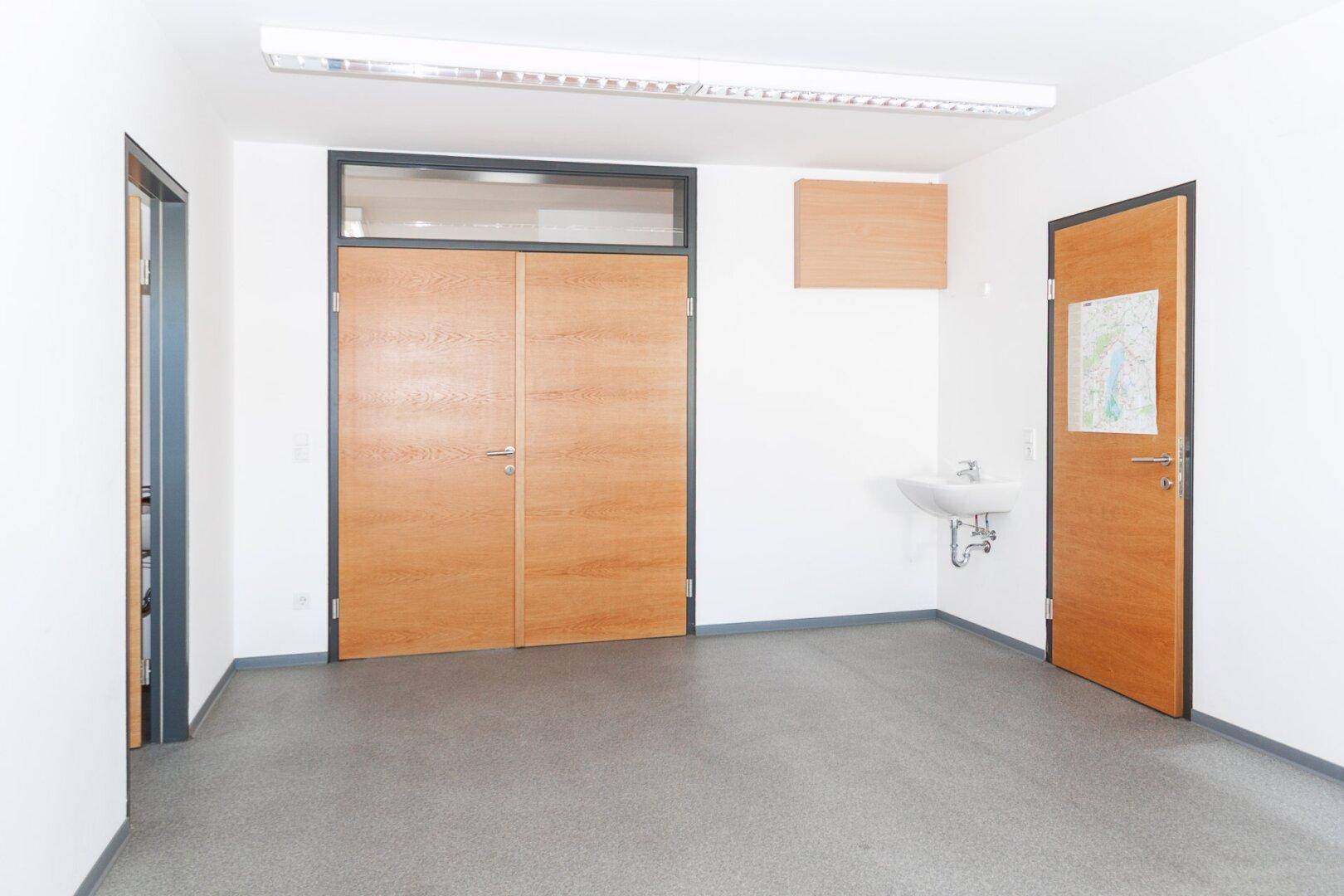 Heller Raum mit Waschbecken und zwei Holztüren, ideal für ein kleines Büro oder eine Teeküche.