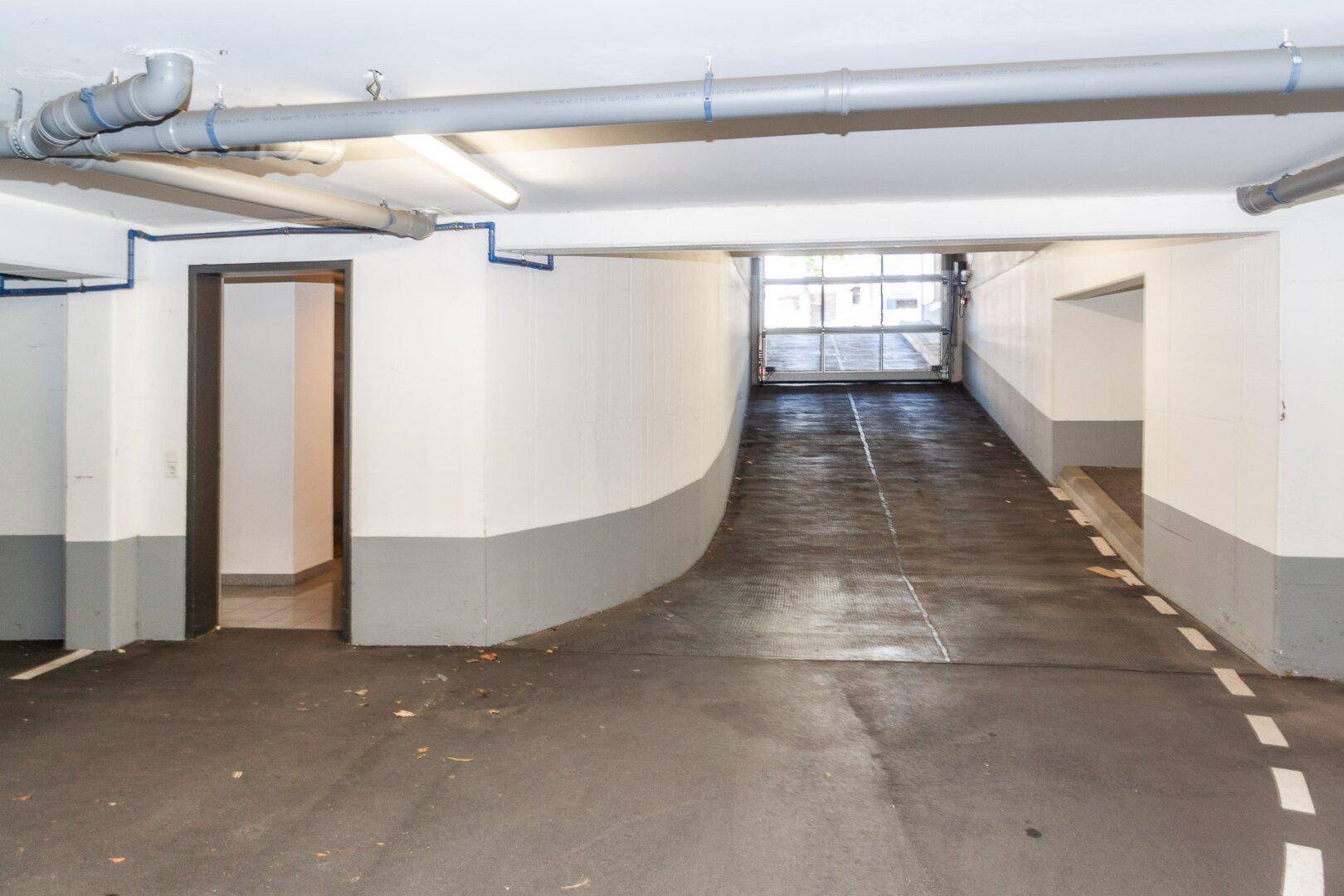 Einfahrt zu einer Tiefgarage mit Rampe und sichtbaren Versorgungsleitungen an der Decke.