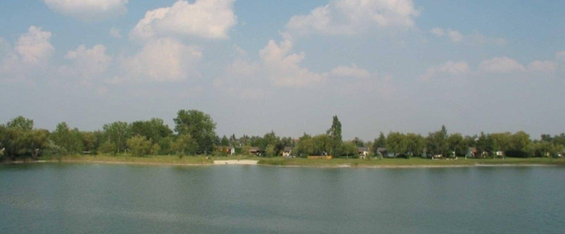 Weitläufiger Blick über einen ruhigen See mit bewaldetem Ufer und vereinzelten Häusern.