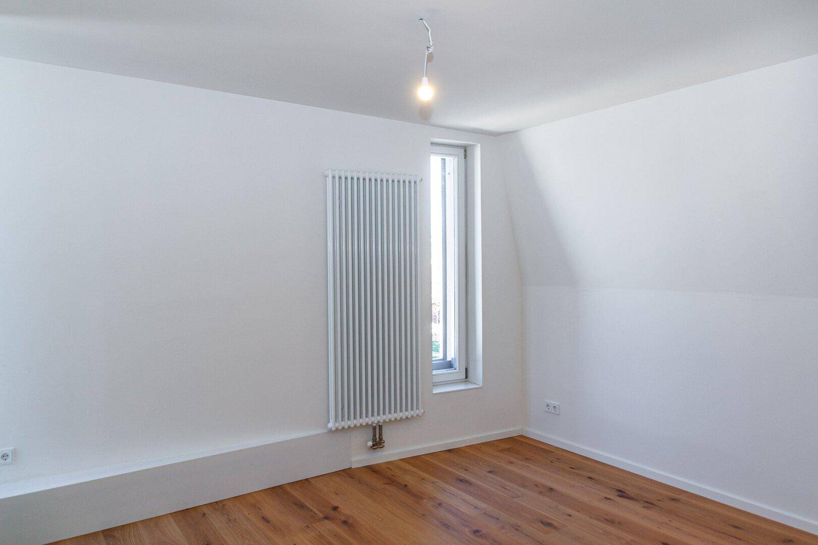 Heller, leerer Raum mit Holzboden, einem hohen Fenster und Heizkörper.