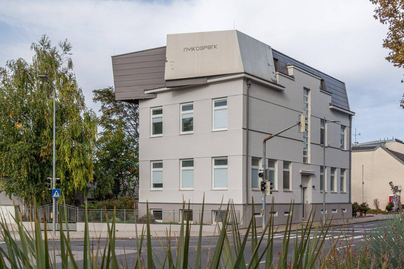 Modernes Gebäude mit heller Fassade, großen Fenstern und markanter Dachkonstruktion an einer Straße.