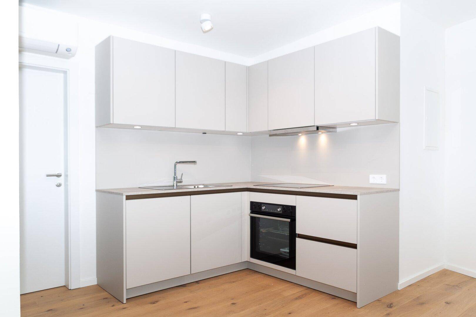 Moderne, kompakte Einbauküche mit hellen Schränken, integriertem Backofen und Holzboden.