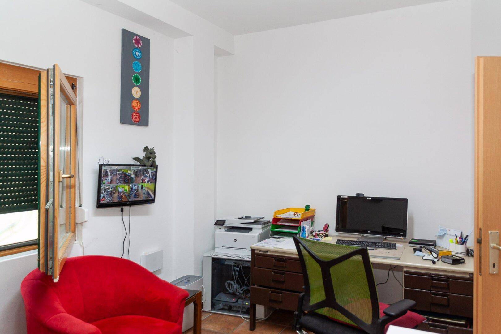 Helles Büro mit Schreibtisch, Computer, rotem Sessel und Fenster mit Rollladen.