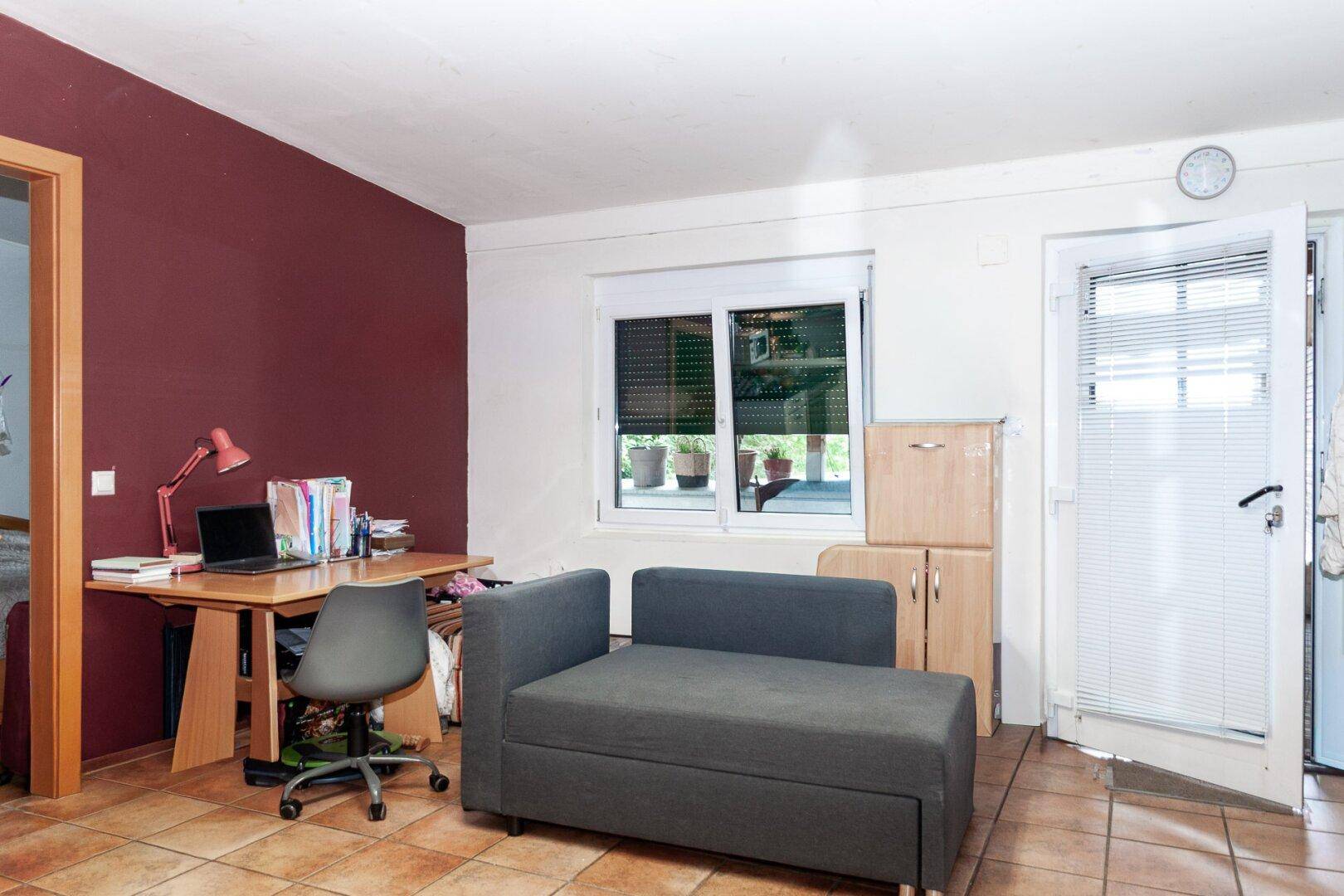Wohnraum mit Schreibtisch, Tagesbett, Fenster mit Rollladen und Zugang nach draußen.