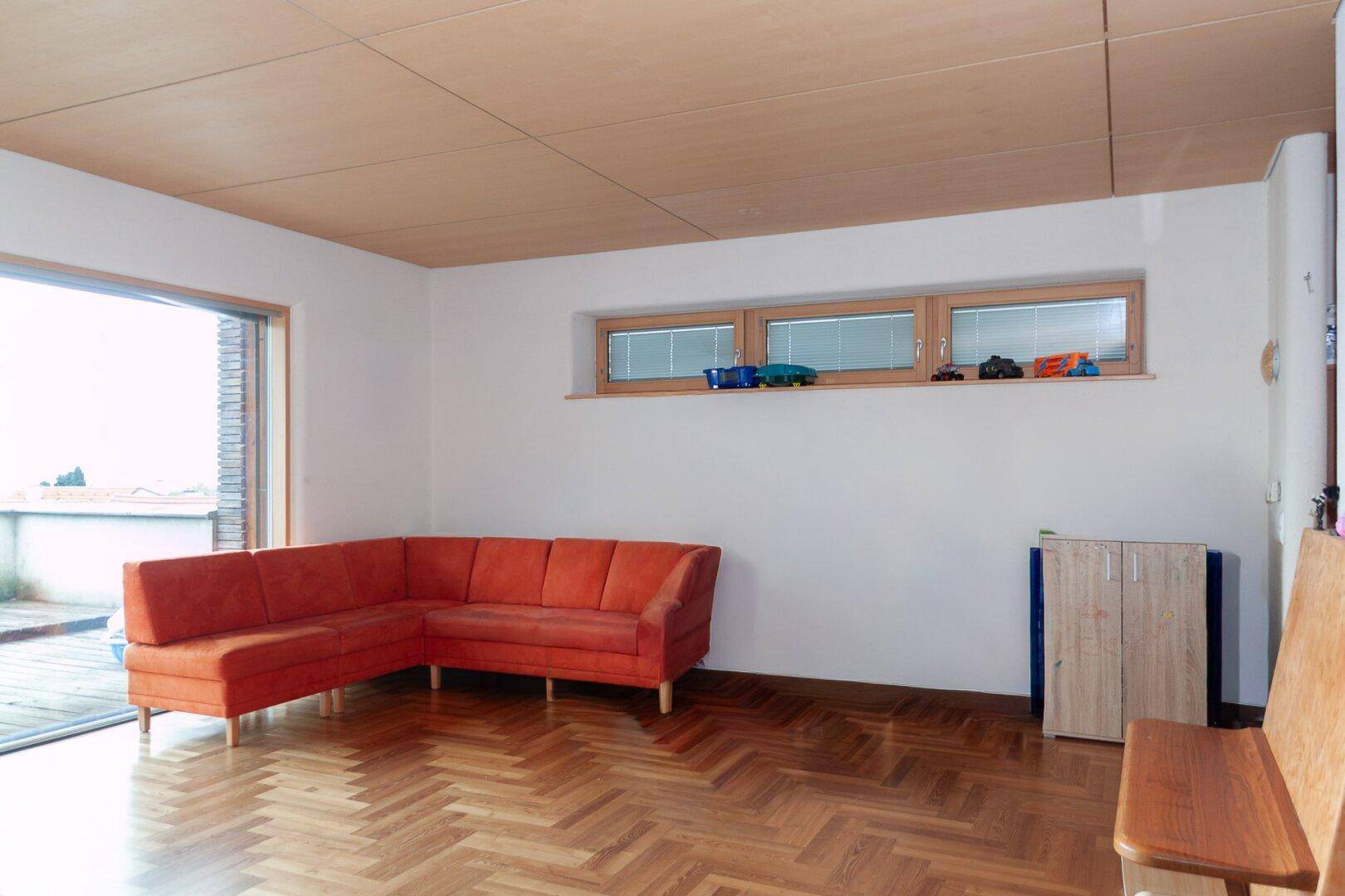 Heller Wohnbereich mit orangefarbenem Sofa, Fischgrätparkett und großen Fenstern.