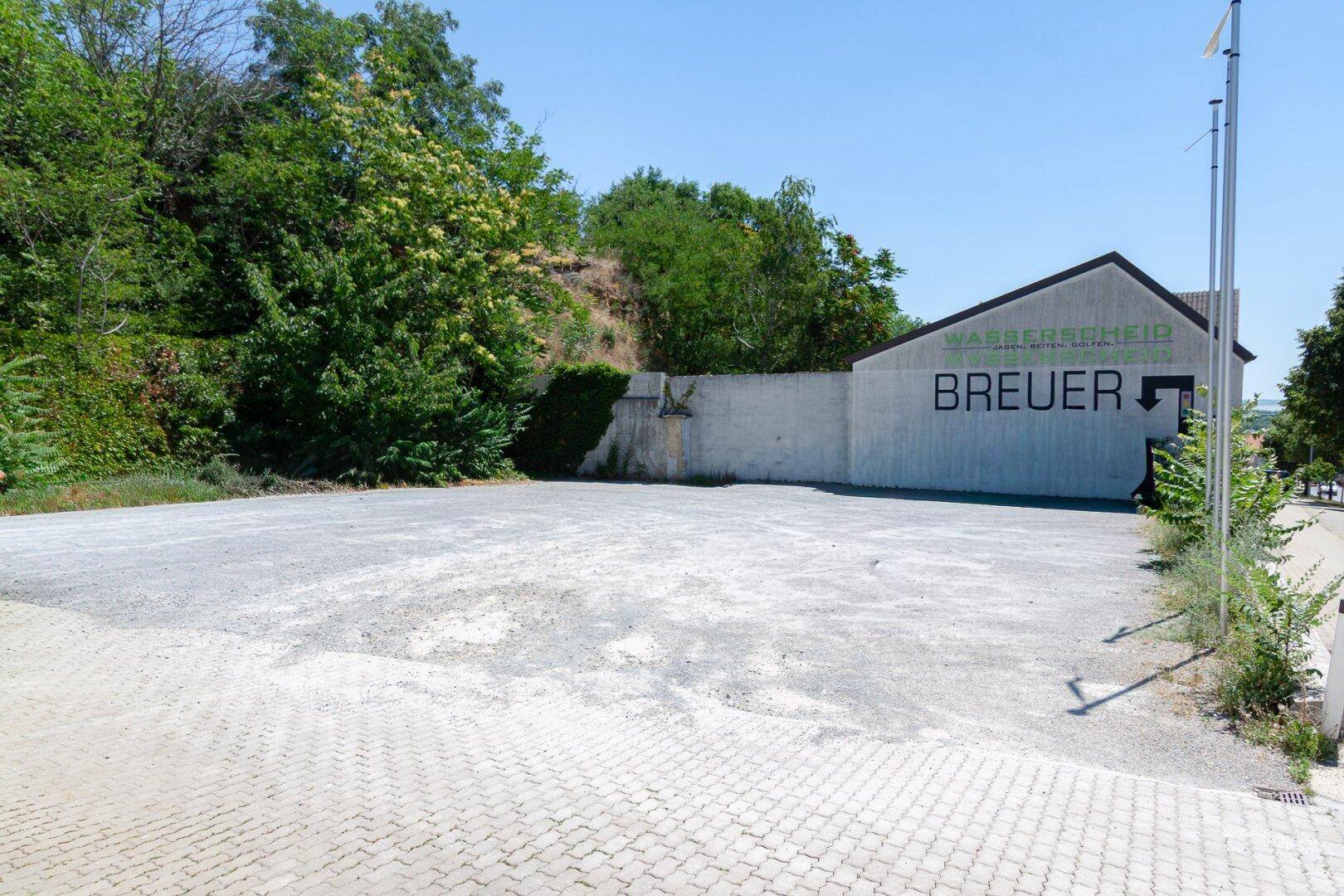 Großer, unbefestigter Parkplatz neben einem Gebäude mit bewachsenem Hang im Hintergrund.