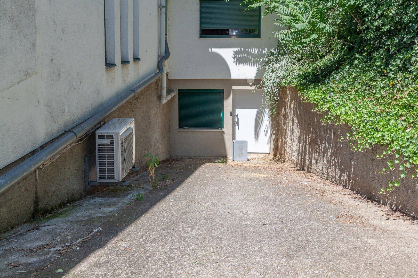 Seitenbereich des Gebäudes mit Klimaanlage, Fensterläden und bewachsenem Hang.