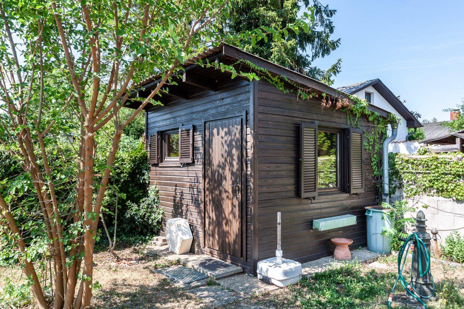 Rustikaler Holzschuppen im Garten, umgeben von Bäumen und Pflanzen an einem sonnigen Tag.