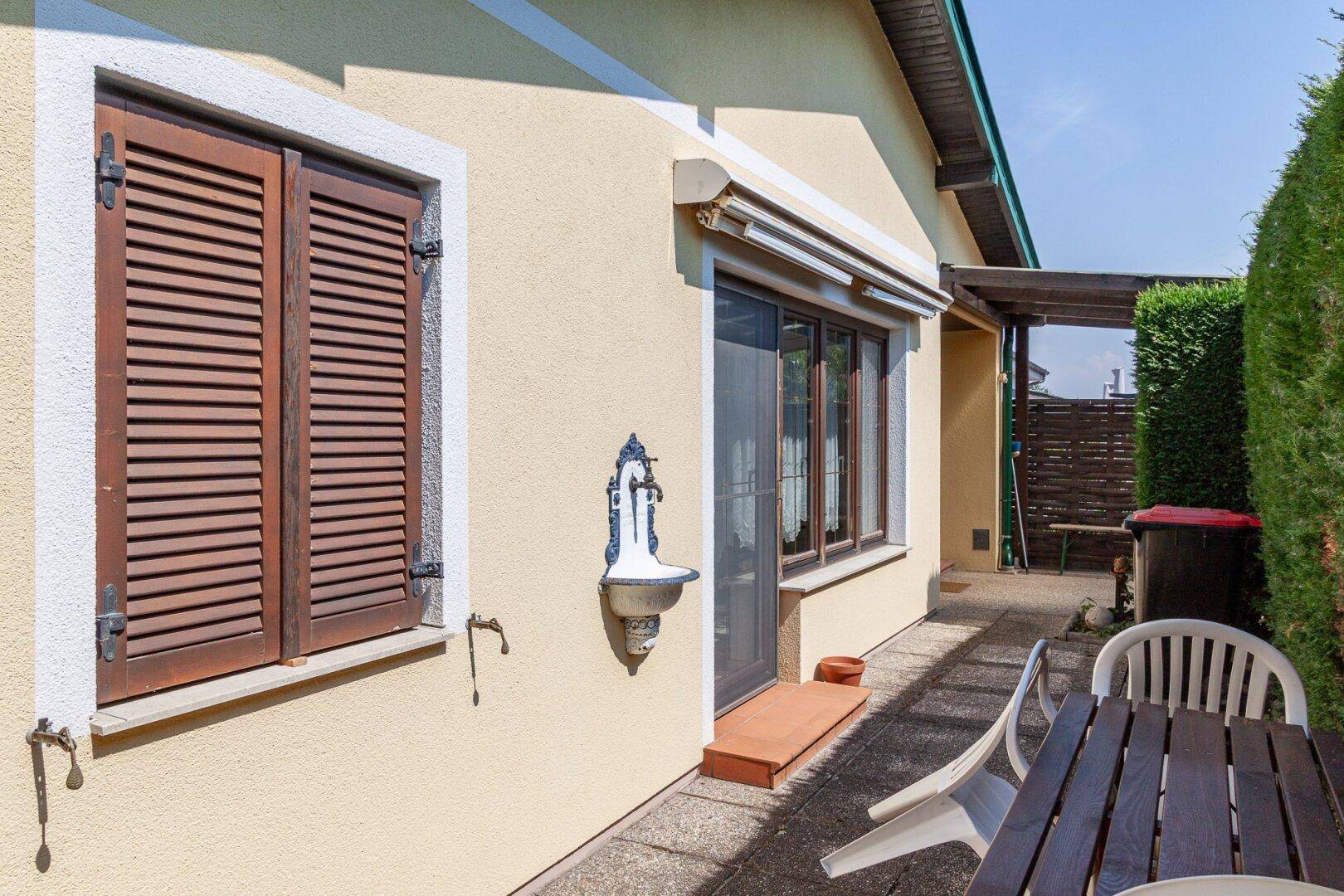 Gepflasterte Terrasse mit Gartenmöbeln, Außenwasserhahn und Fenster mit Holzläden.
