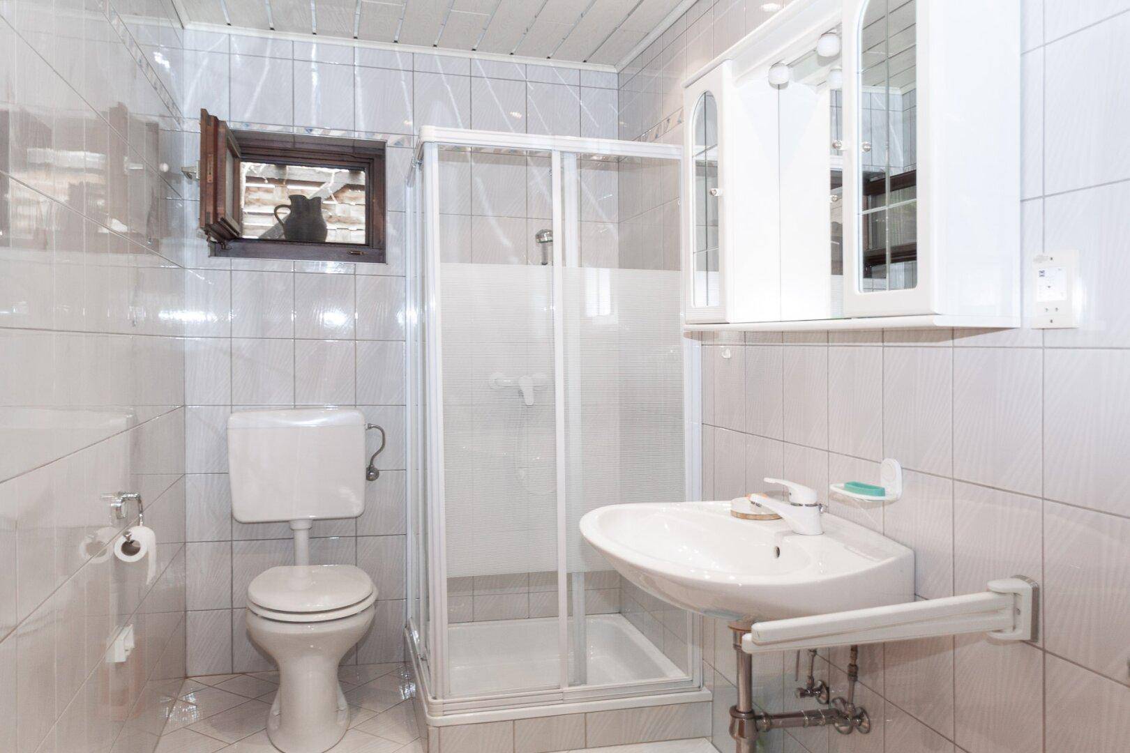Badezimmer mit weißen Fliesen, Toilette, Waschbecken und Duschkabine, sowie einem kleinen Fenster.