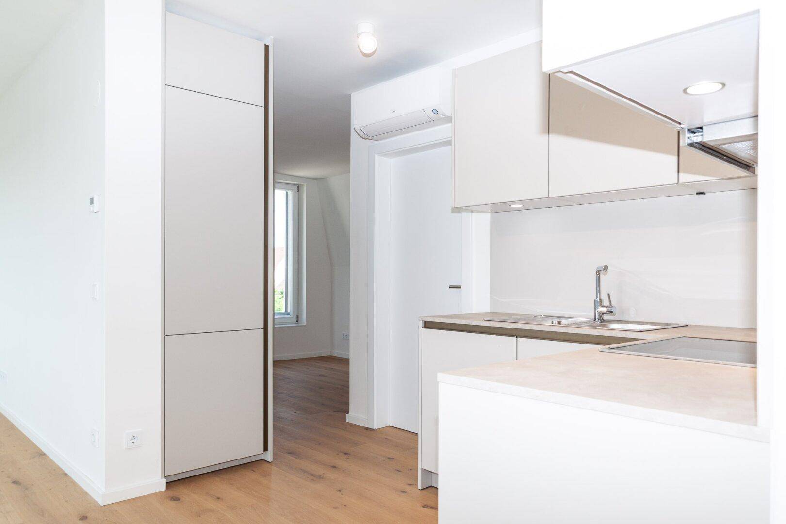 Helle Küchenzeile mit integriertem Kühlschrank und Holzboden, offen zum angrenzenden Wohnbereich.