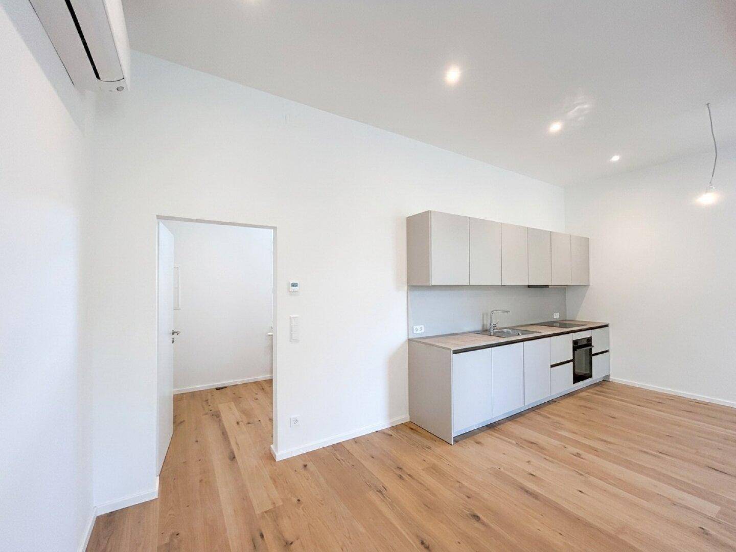 Offene Küche in hellem Raum mit Holzboden, Klimaanlage und Zugang zu einem weiteren Zimmer.