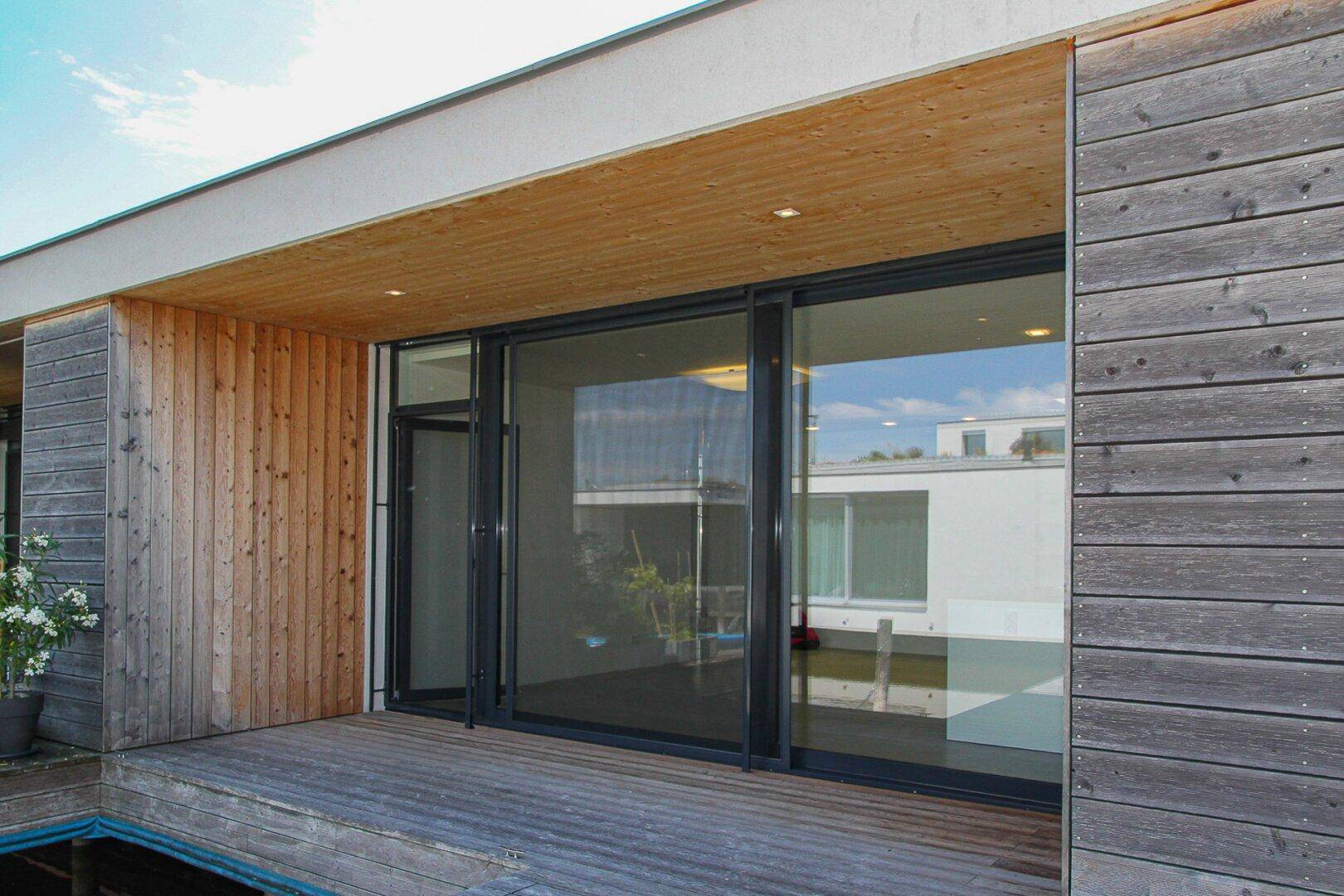 Holzverkleidete Fassade mit großen Glasschiebetüren, die zu einer Holzterrasse am Wasser führen.