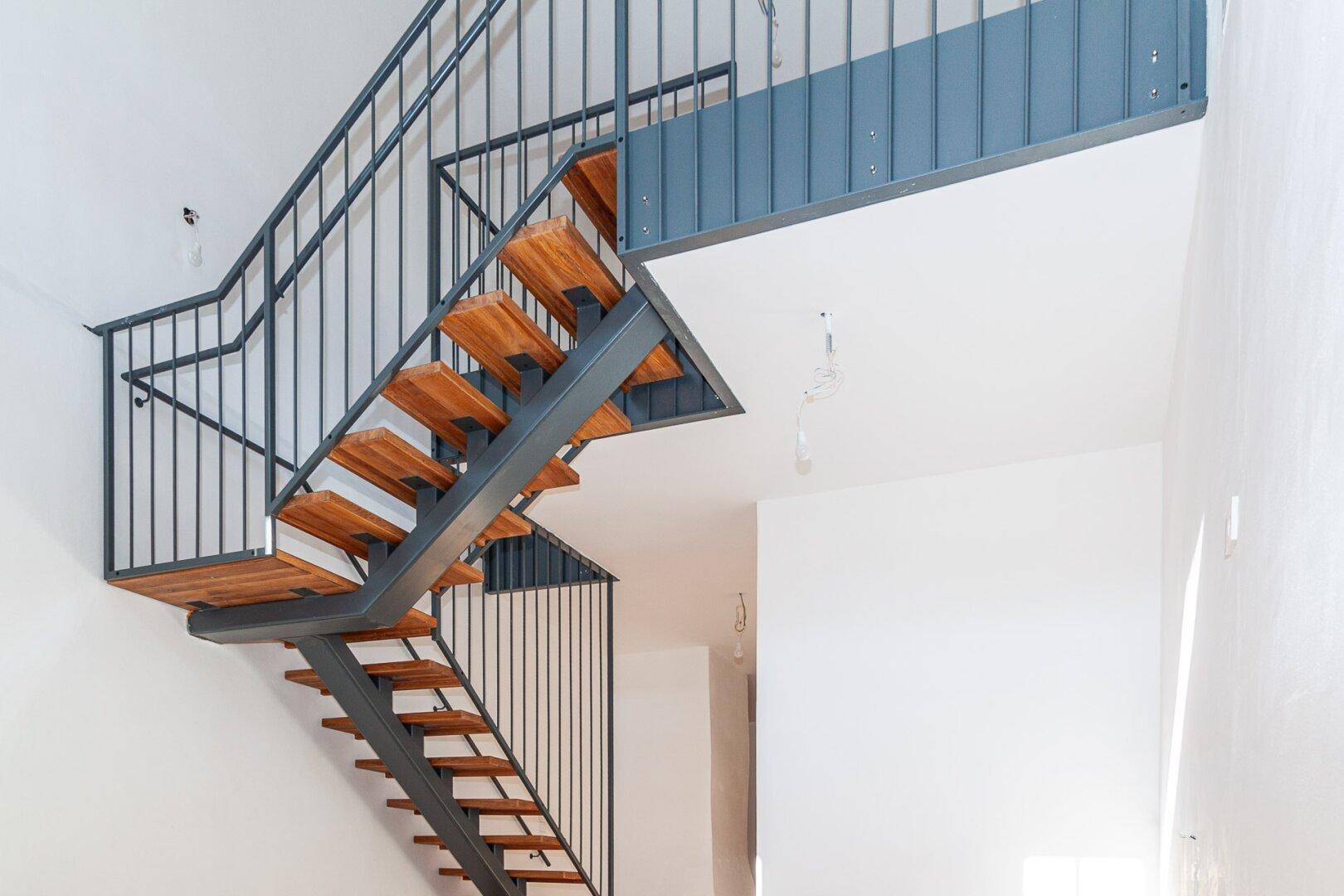 Mehrstufige Holztreppe mit schwarzem Metallgeländer in einem hohen, hellen Innenraum mit viel Licht.