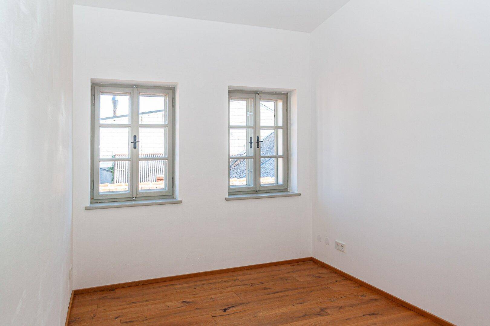 Helles Schlafzimmer mit Holzboden und zwei Fenstern, die viel Tageslicht hereinlassen.