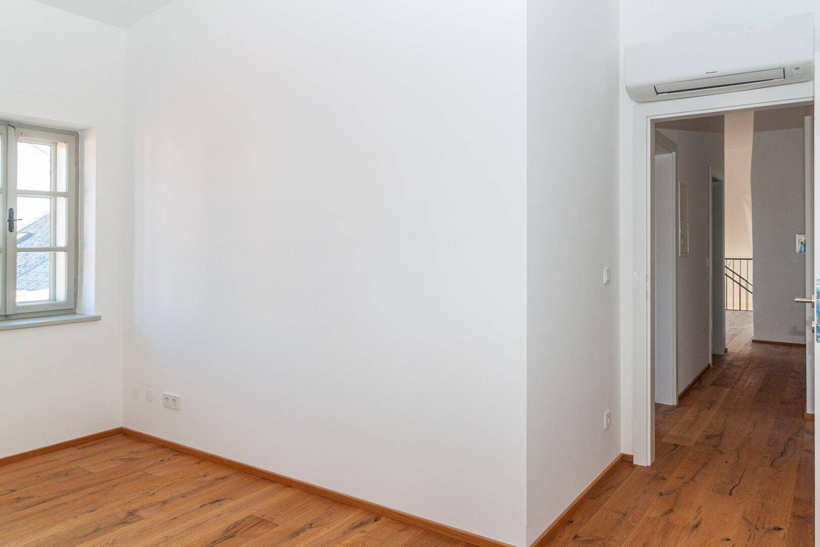 Helles Schlafzimmer mit Holzboden und Fenster, verbunden mit einem Flur mit Klimaanlage.