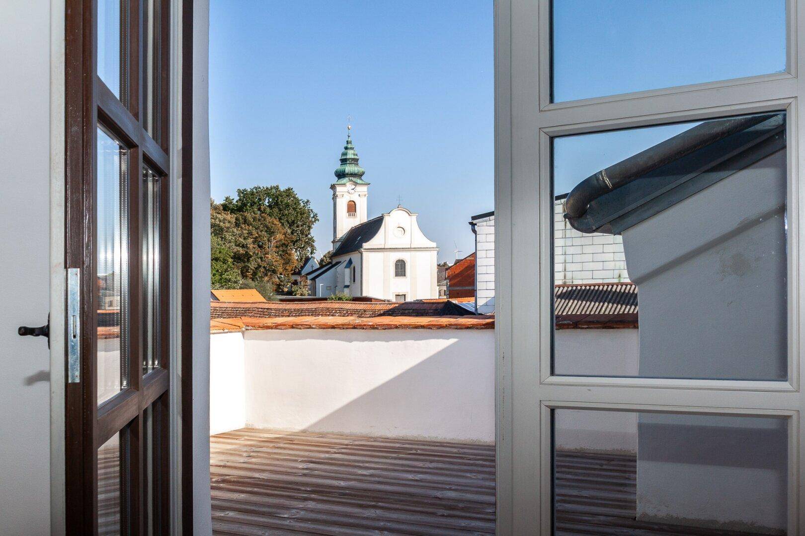 Blick von der Terrasse auf eine historische Kirche und die umliegende Dachlandschaft bei blauem Himmel.