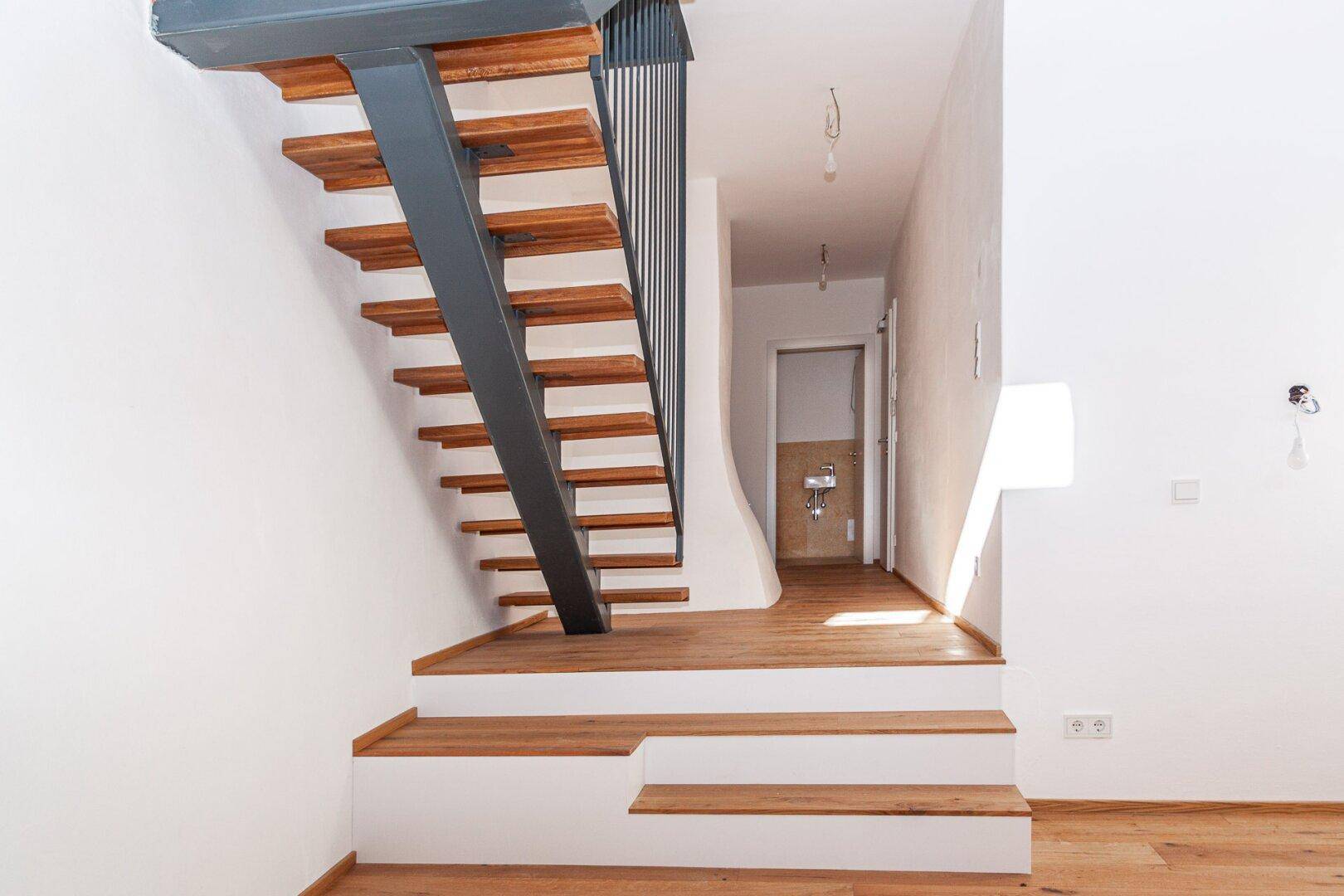 Moderne Holztreppe mit Metallgeländer führt in einen hellen Flur mit Holzboden und hohen Decken.