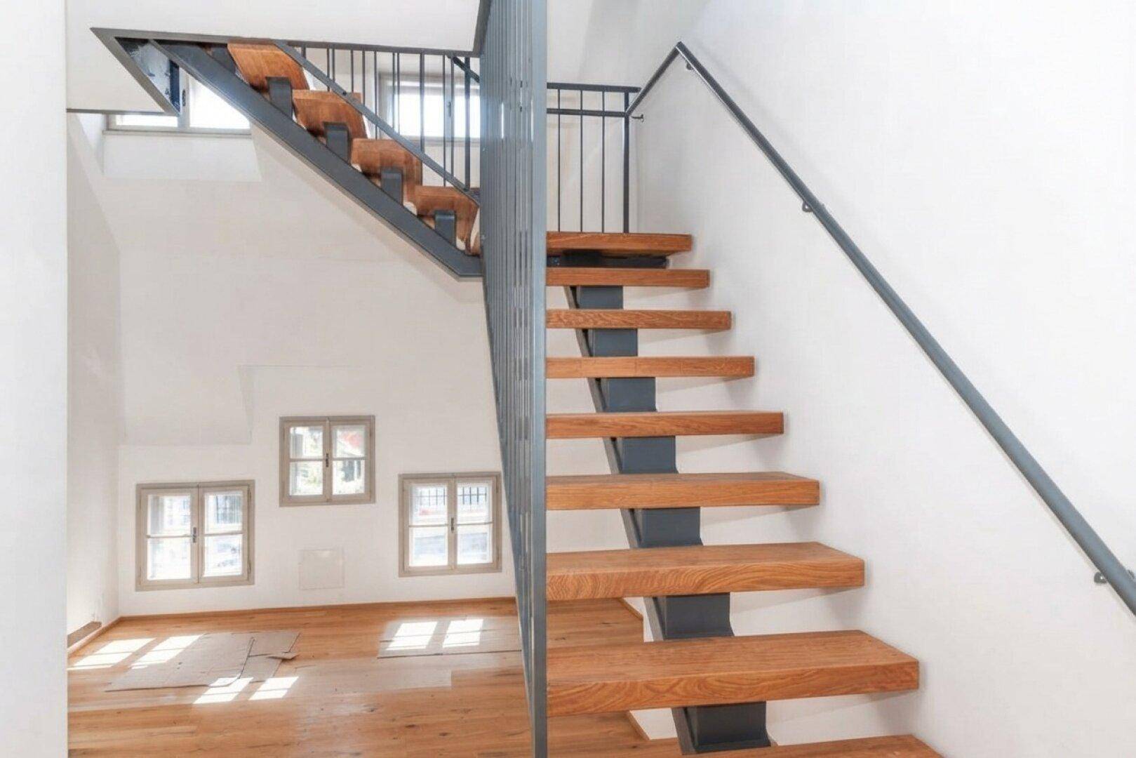 Offene Holztreppe mit Metallgeländer in einem hellen Raum mit mehreren Fenstern und Holzboden.