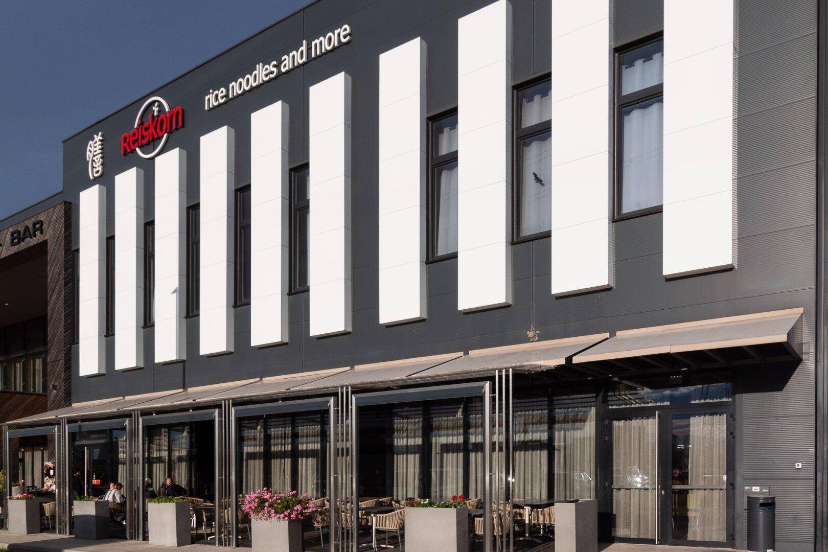 Moderne Restaurantfassade mit Außenterrasse, Markisen und Sitzgelegenheiten unter freiem Himmel.