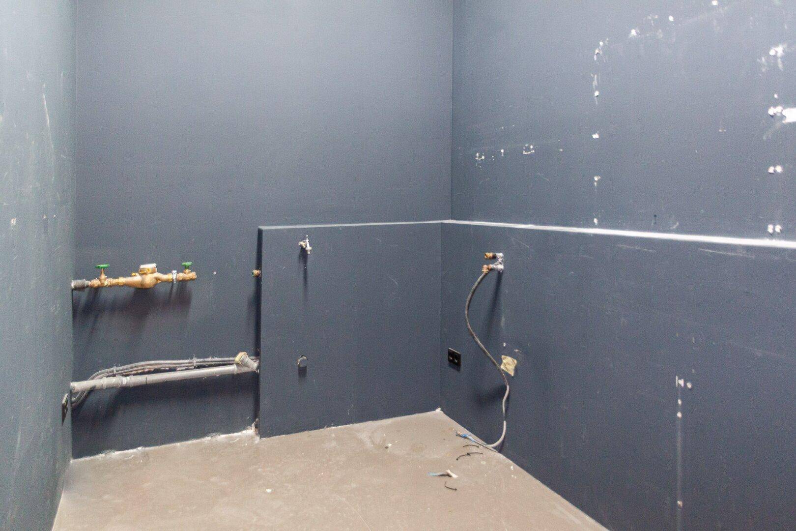 Unfertiger Versorgungsraum mit grauen Wänden und sichtbaren Wasserleitungen und Anschlüssen.