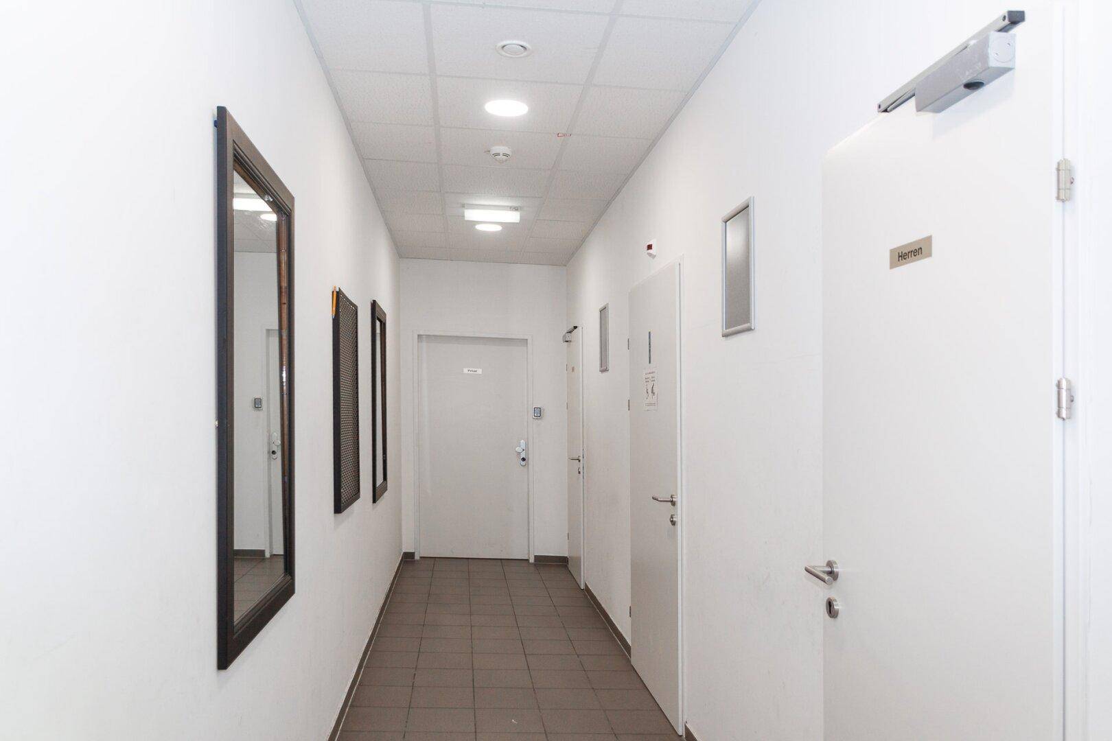Heller Flur mit weißen Wänden, braunen Fliesen und mehreren Türen zu verschiedenen Räumen.