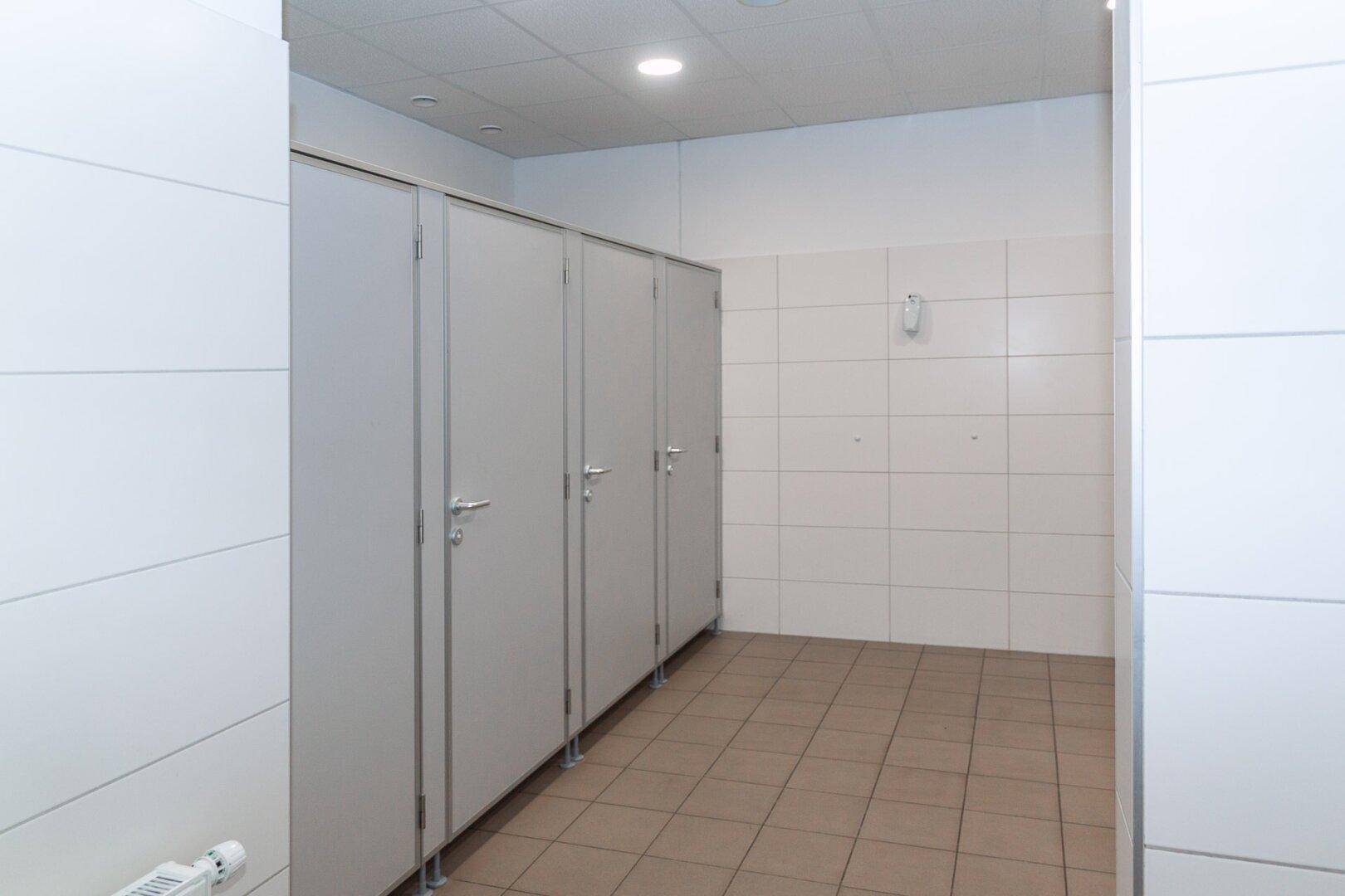 Toilettenbereich mit mehreren Kabinen, weißen Fliesen und braunem Fliesenboden.