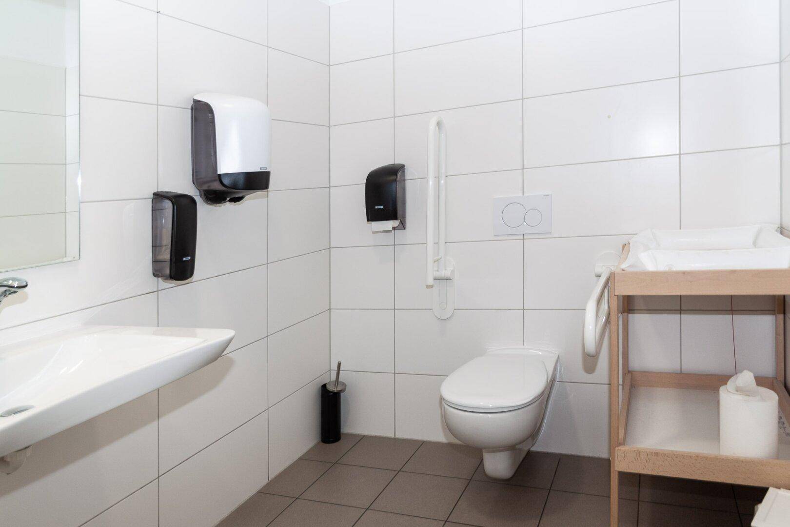 Barrierefreier Toilettenraum mit Wickeltisch, Haltegriffen und wandhängendem WC.