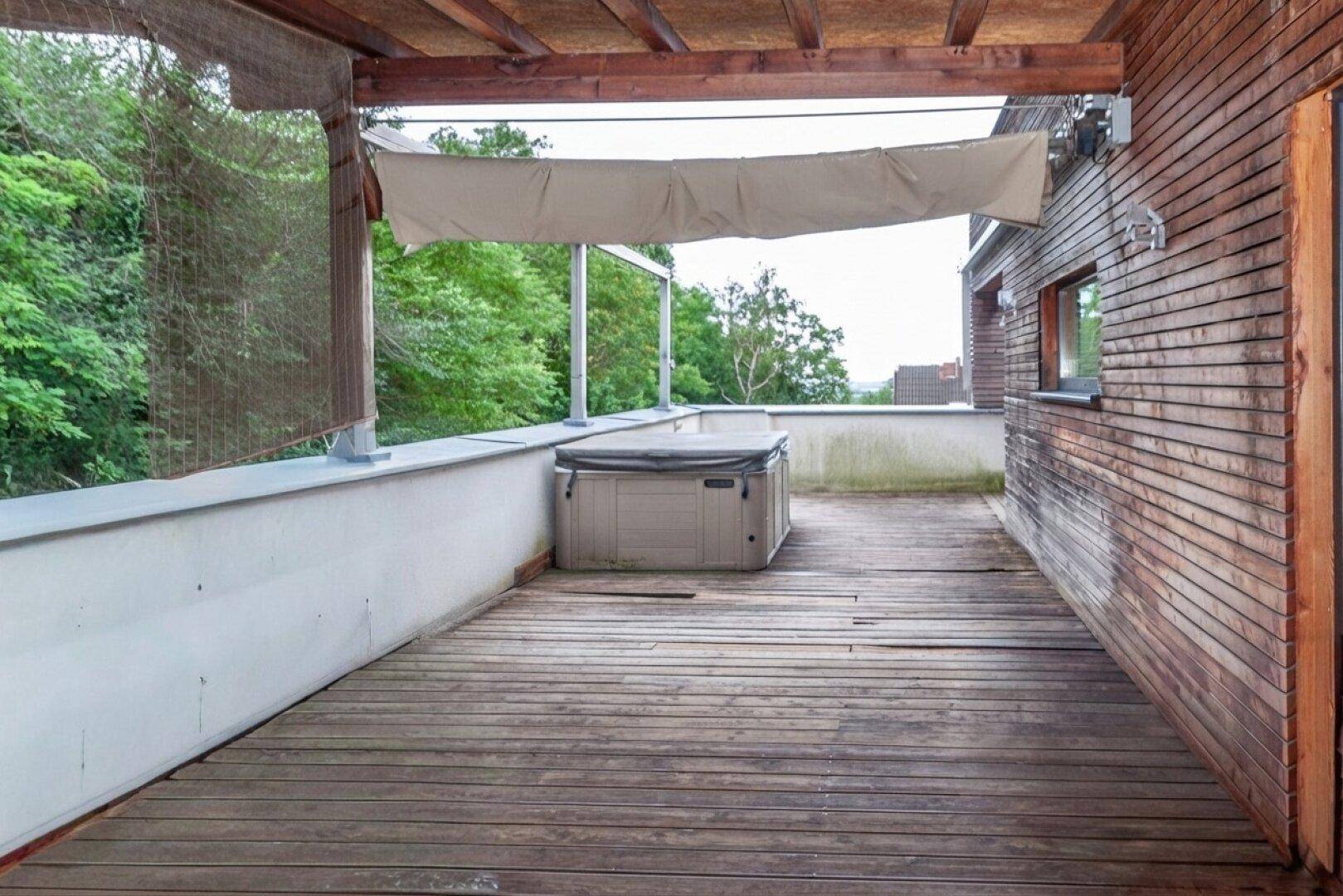 Große Holzterrasse mit Whirlpool und einer Überdachung, umgeben von viel Grün.