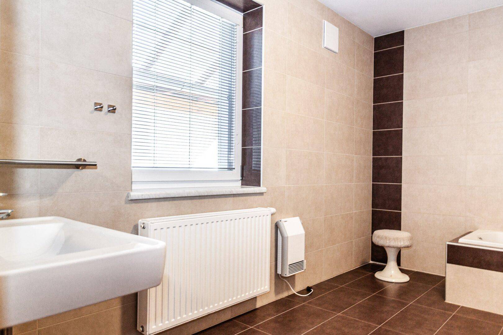 Badezimmer mit beigefarbenen Fliesen, Waschbecken, Heizkörper und Fenster mit Jalousien.
