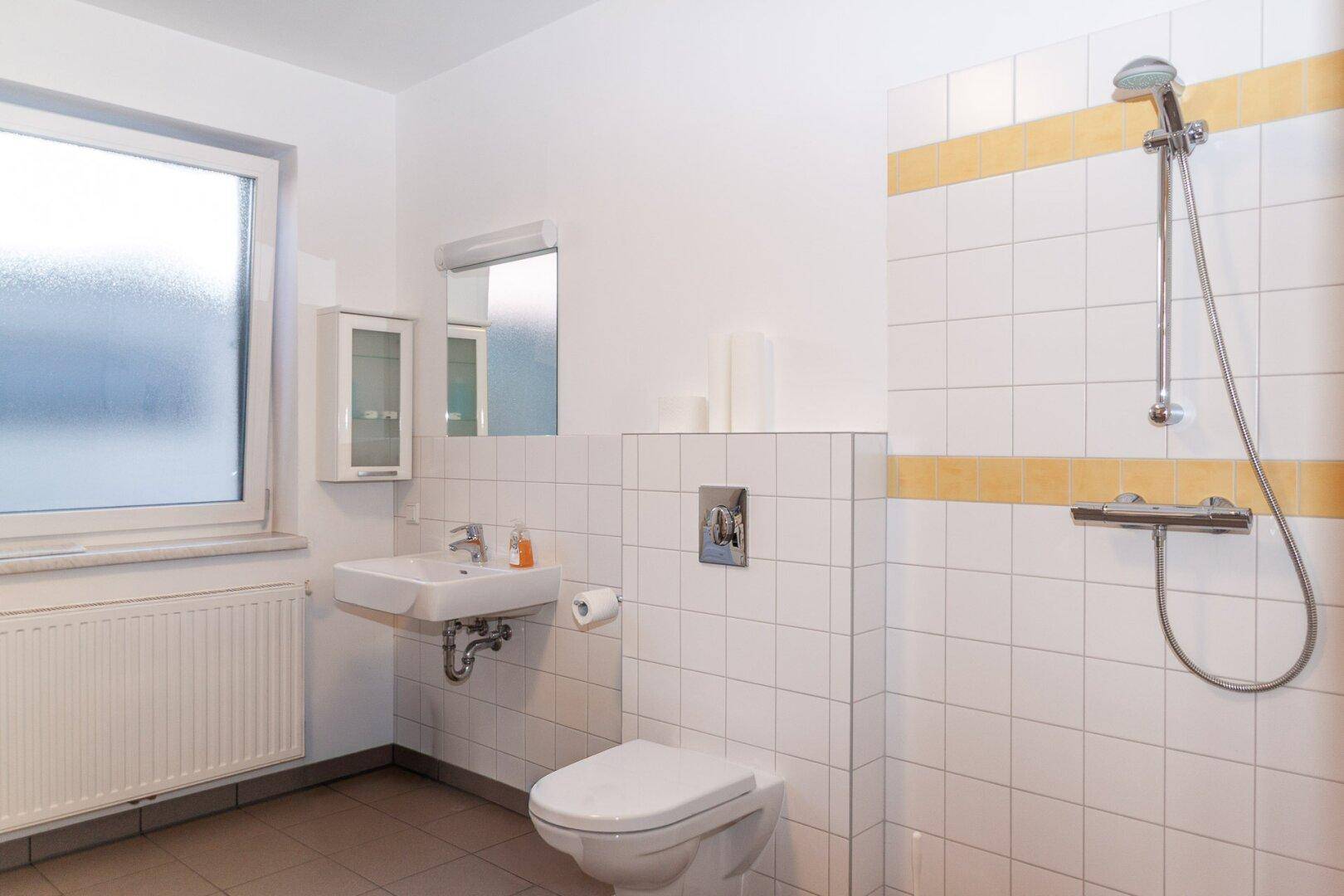 Helles Badezimmer mit weiß-gelben Fliesen, Dusche, WC und Fenster für Tageslicht.