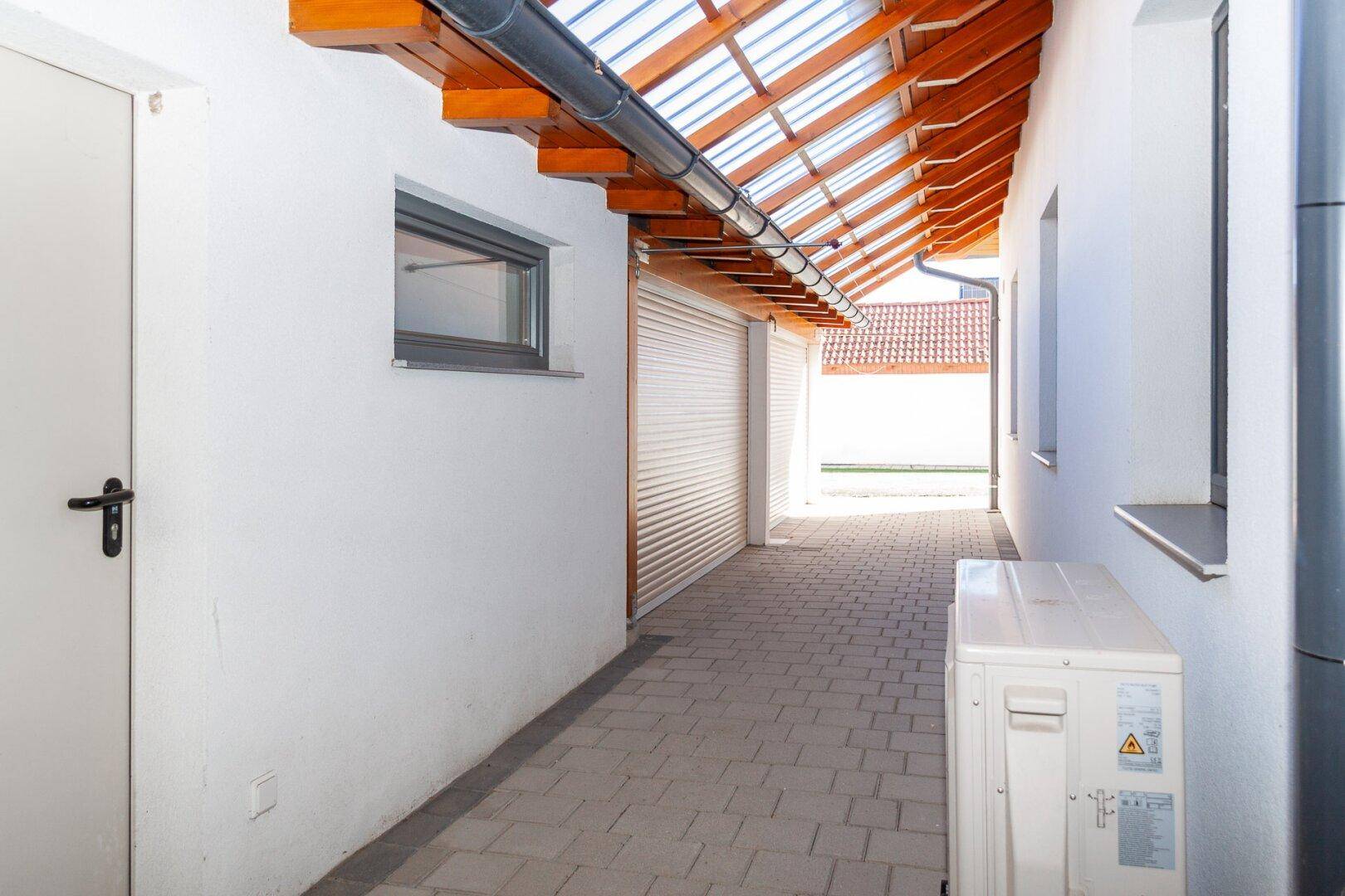 Überdachter Gang mit gepflastertem Boden und transparentem Dach, führt zur Garage.