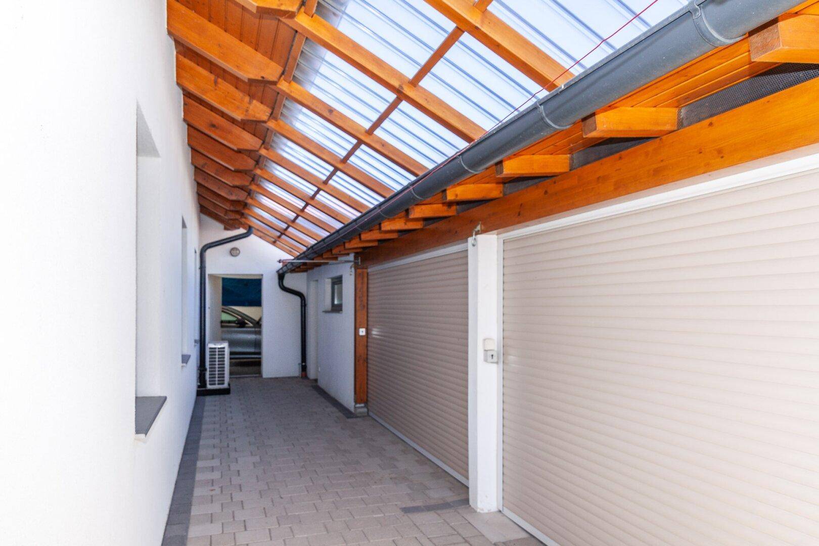 Überdachter Bereich mit Holzbalken und transparentem Dach, führt zu Garagentoren.