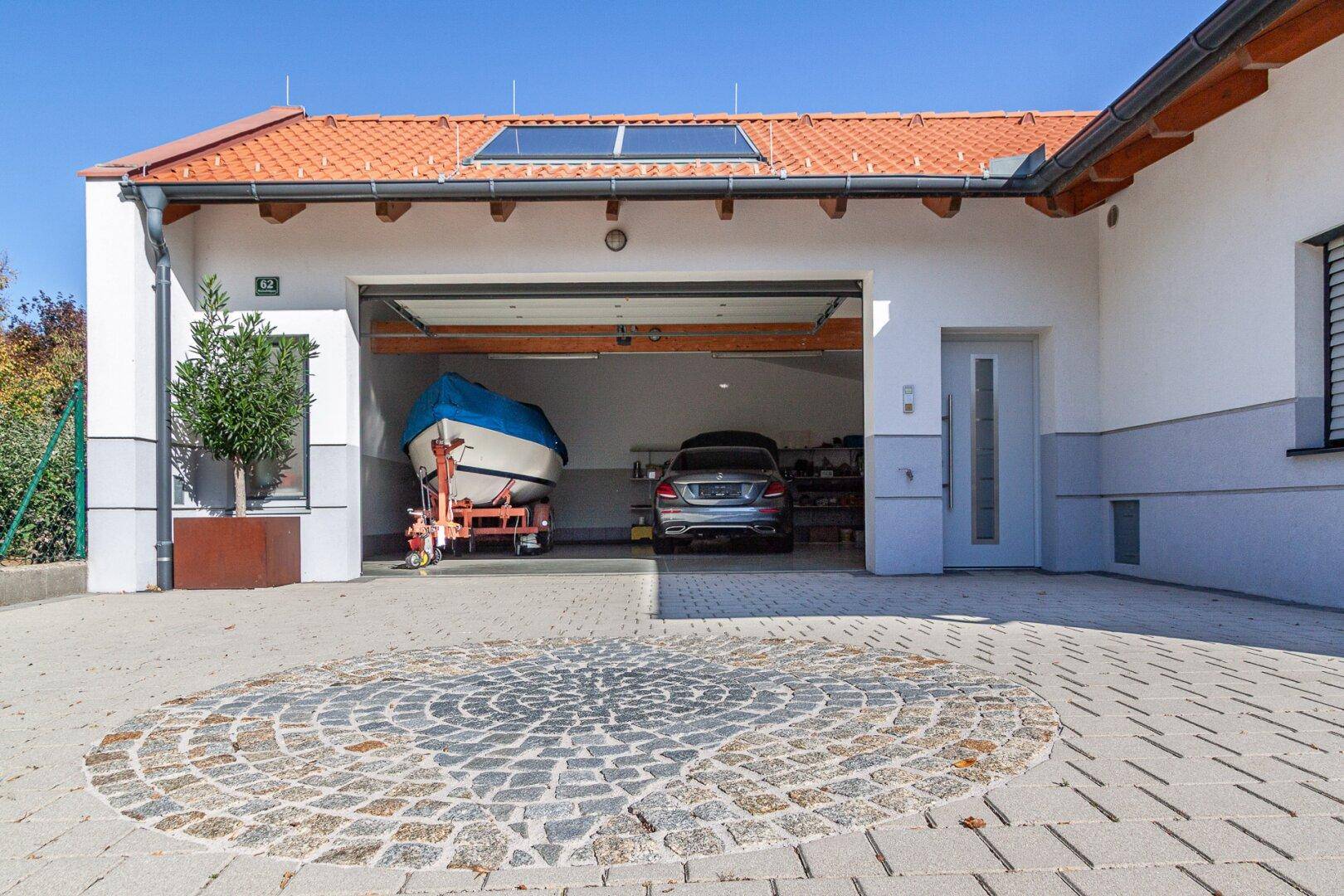 Offene Garage mit geparktem Auto und Boot, davor eine gepflasterte Auffahrt mit Mosaik.