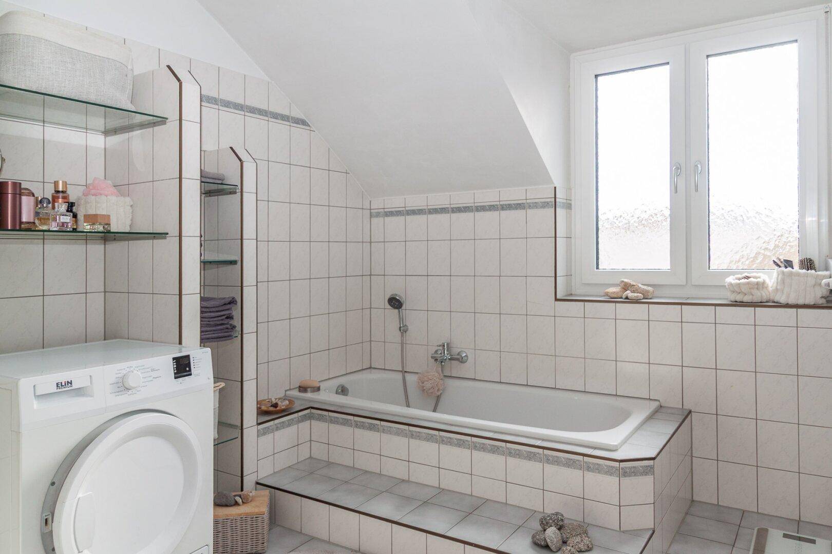 Helles Badezimmer mit Badewanne, Fliesenboden und Dachschräge, inklusive Waschmaschine.