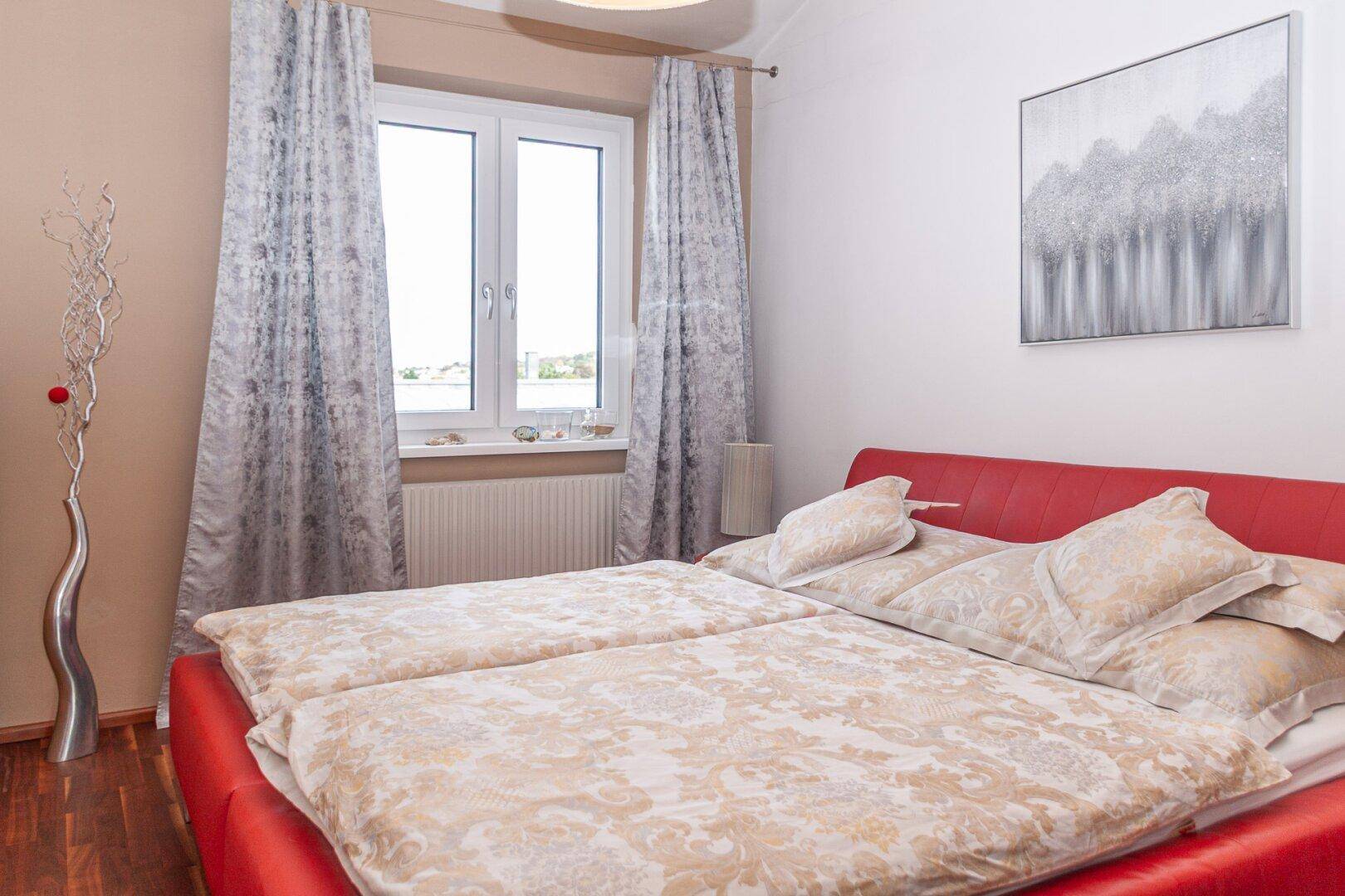 Schlafzimmer mit rotem Bett, Parkettboden und Fenster mit Blick nach draußen.