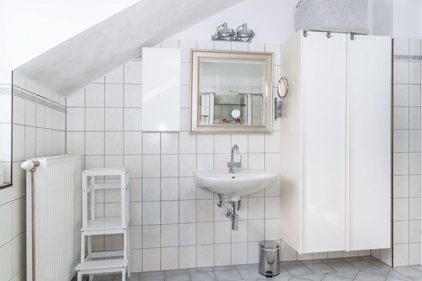 Badezimmer mit Waschbecken, Spiegel und weißen Fliesen unter der Dachschräge.