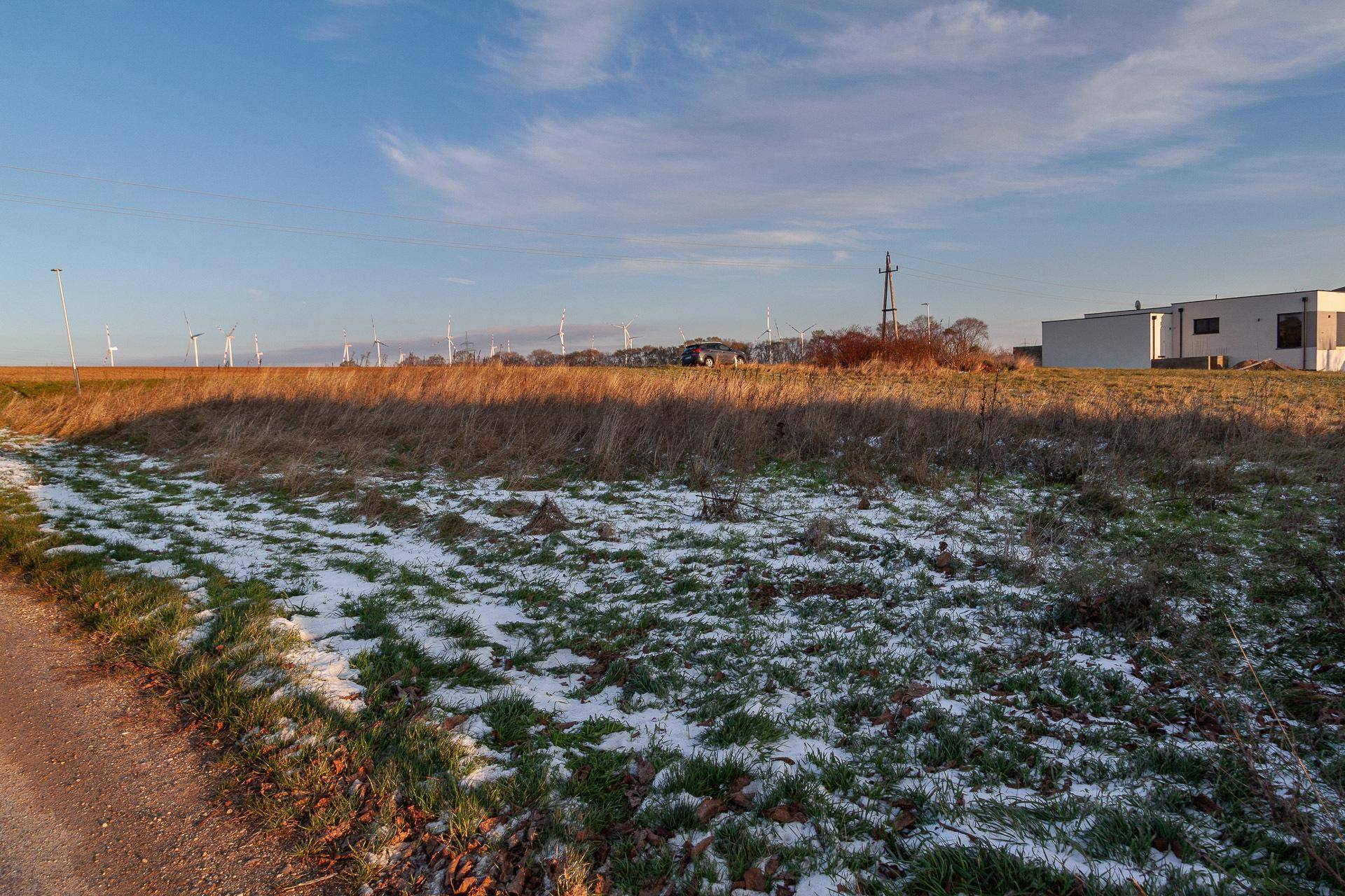 Offene Landschaft mit Feldern, leichtem Schneefall und Windkraftanlagen in der Ferne unter klarem Himmel.