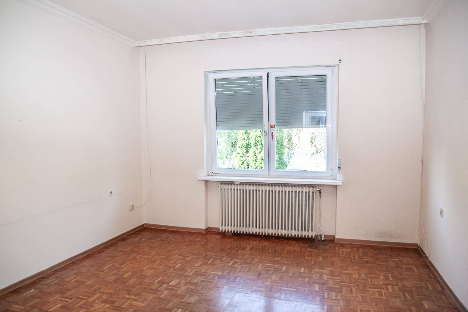 Schlafzimmer mit Parkettboden, großem Fenster und Heizkörper, bereit zur Renovierung.