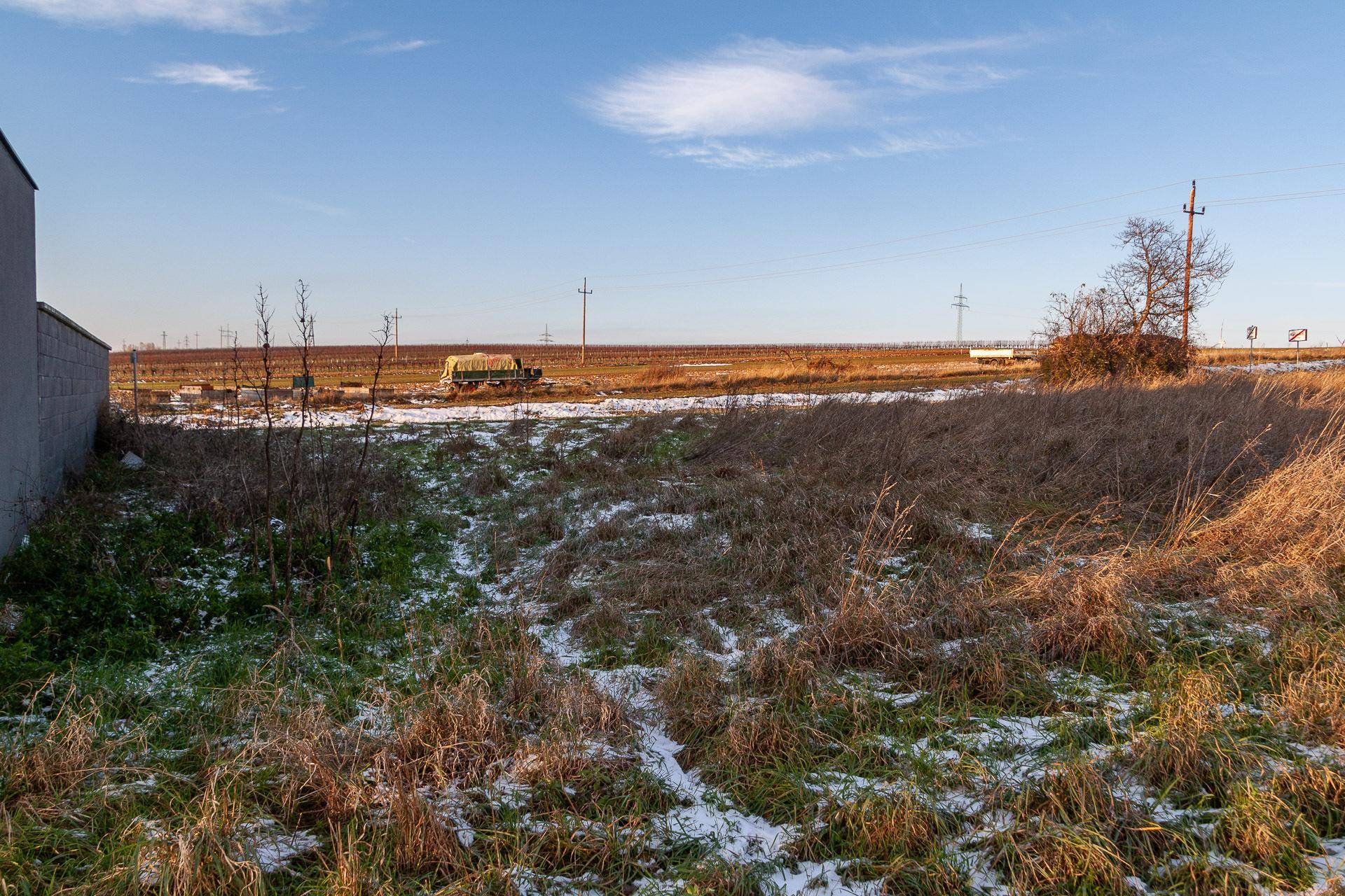 Blick über ein trockenes Feld mit Schneeresten neben einem Gebäude in ländlicher Umgebung.