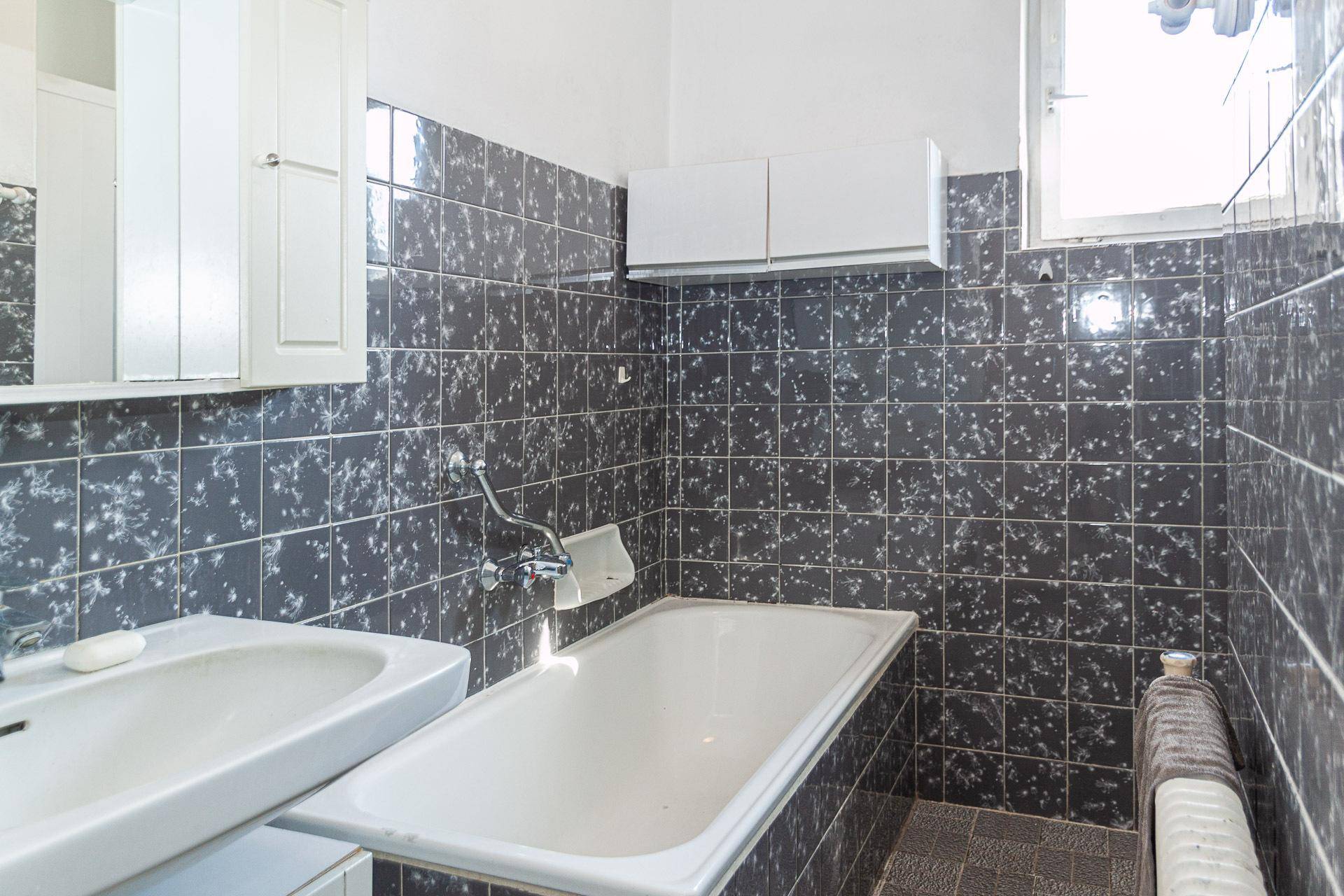 Badezimmer mit Badewanne, Waschbecken und dunklen Fliesen, ausgestattet mit einem Fenster.