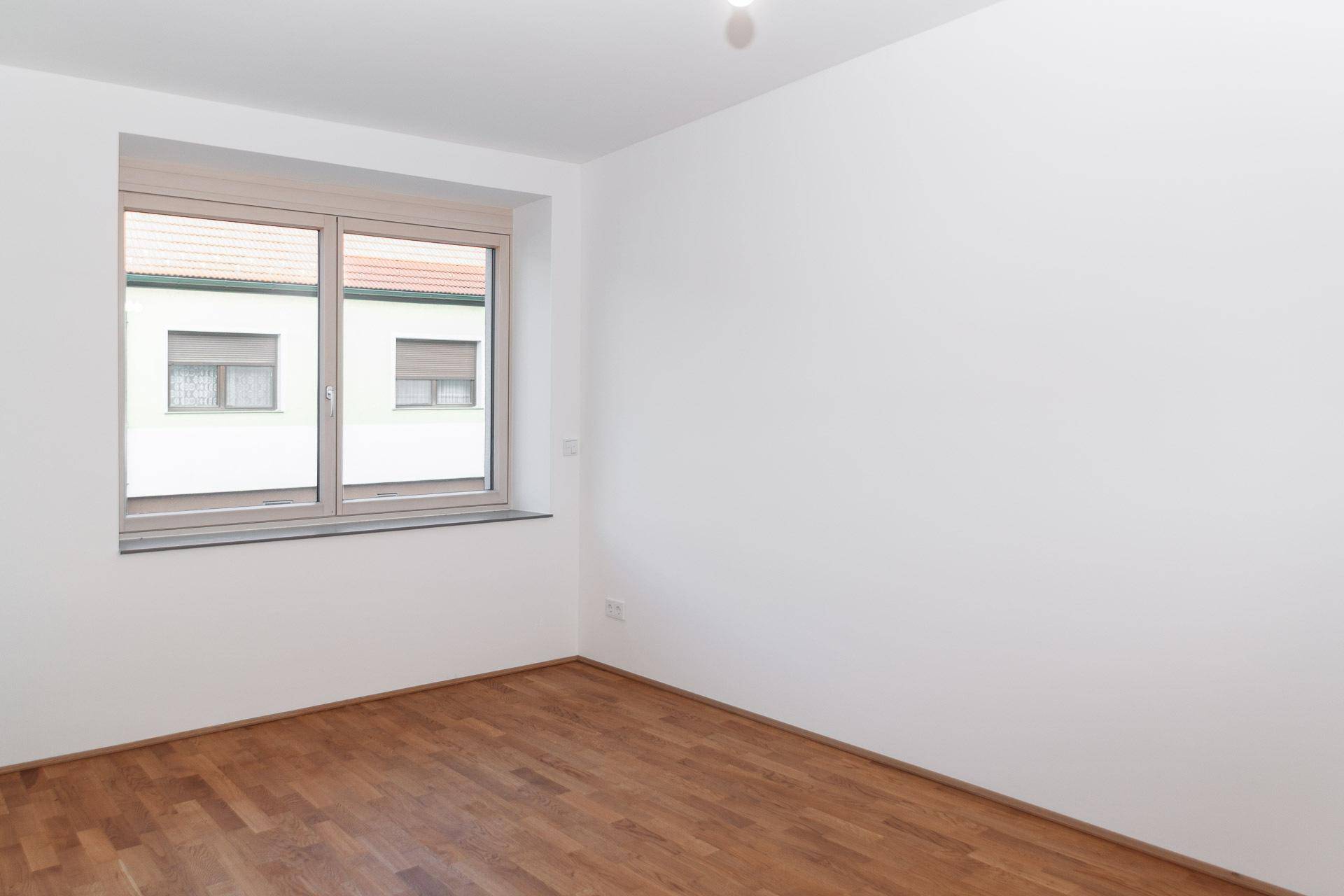 Leeres Schlafzimmer mit Holzboden und Fenster, das viel Tageslicht hereinlässt.