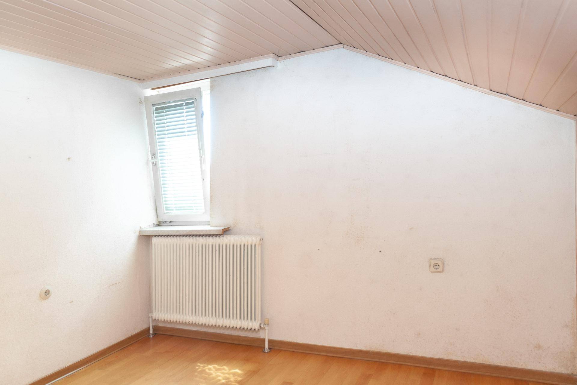 Dachzimmer mit schräger Decke, hellem Boden und Fenster, das viel Licht hereinlässt.