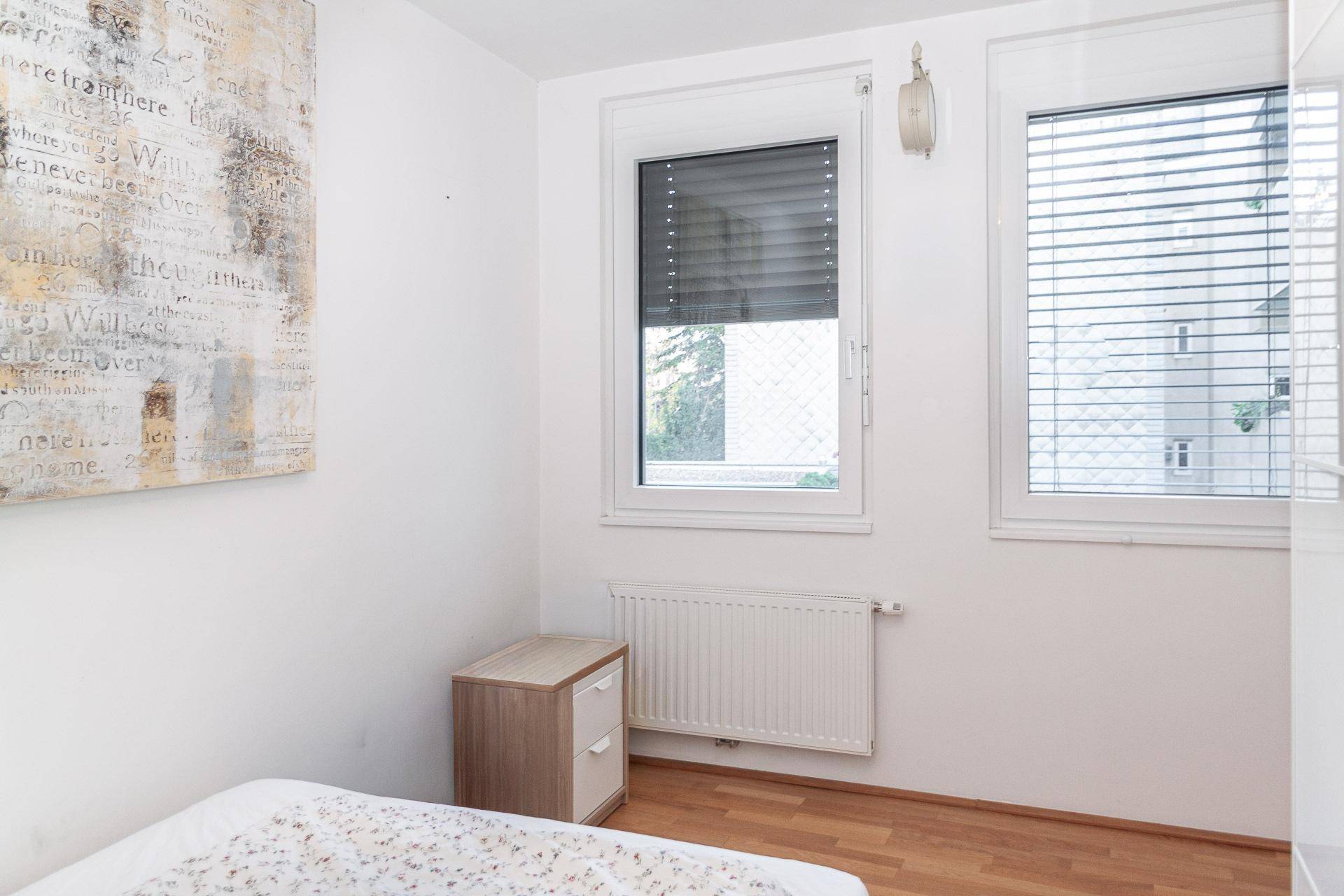 Helles Schlafzimmer mit Holzboden, Fenster mit Jalousien und einfacher Einrichtung.