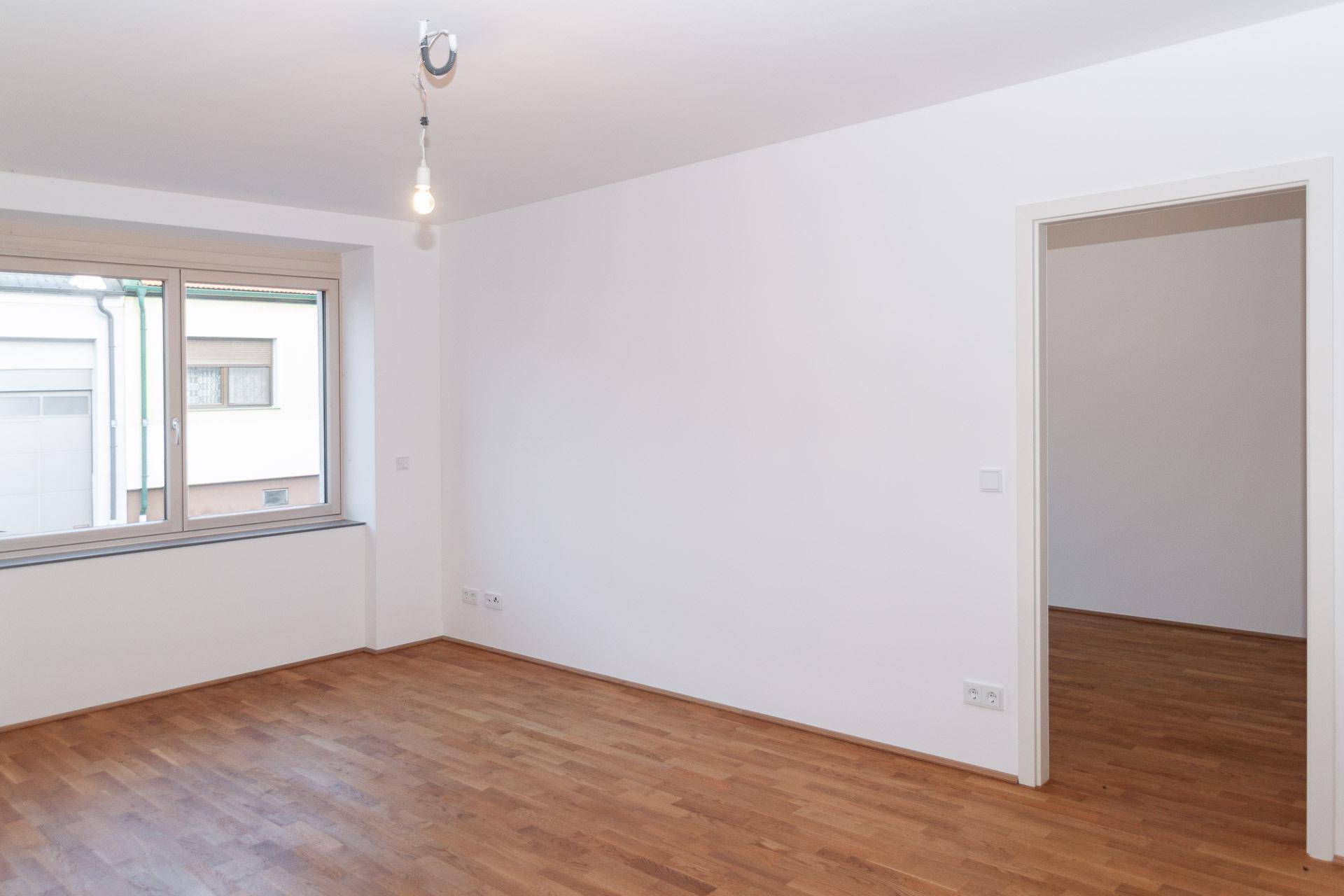 Heller, leerer Raum mit Holzboden und großem Fenster, ideal für Wohn- oder Schlafzimmer.