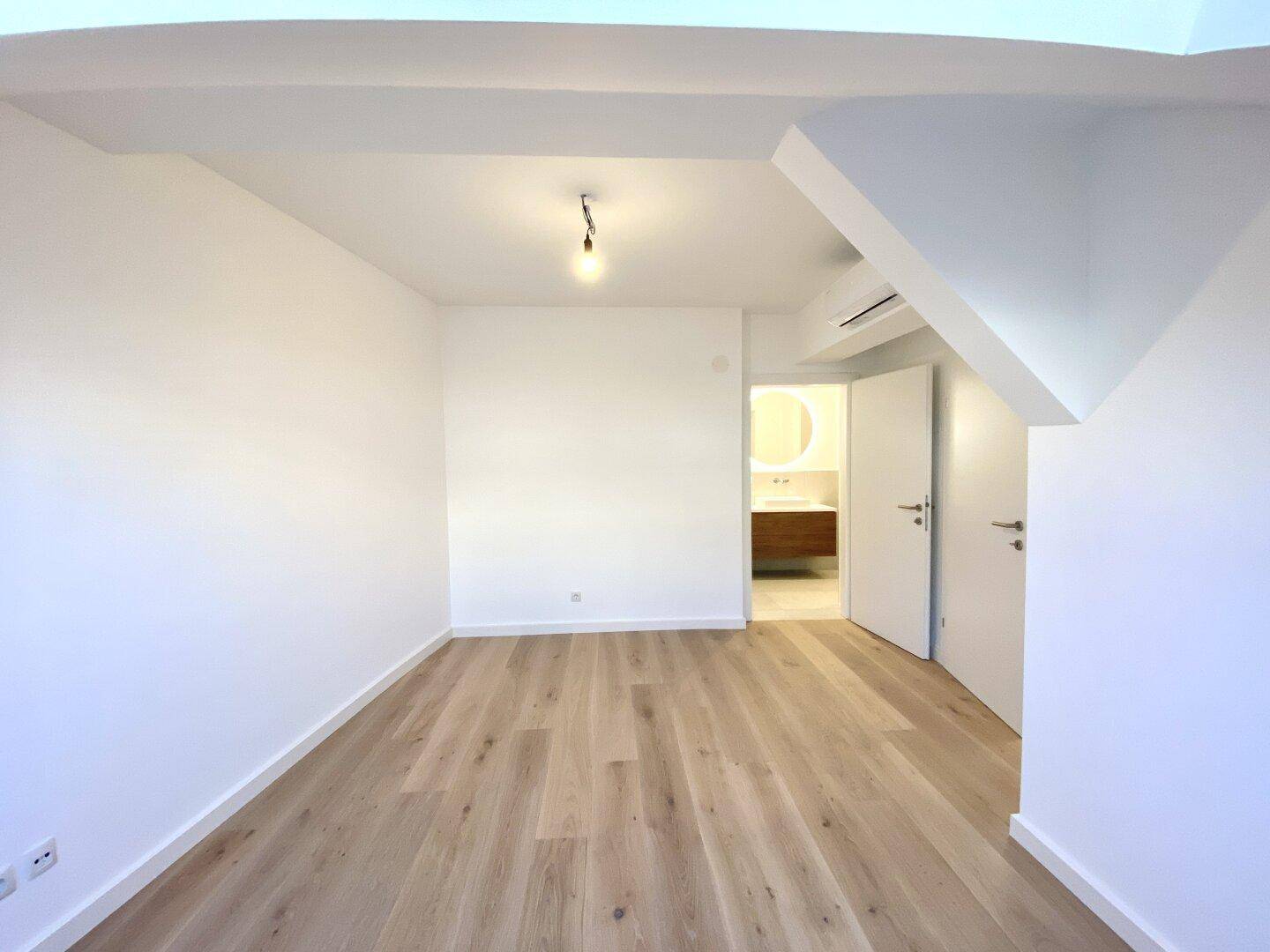 Leerer Raum mit Holzboden und Zugang zu einem modernen Badezimmer, ideal als Schlafzimmer.