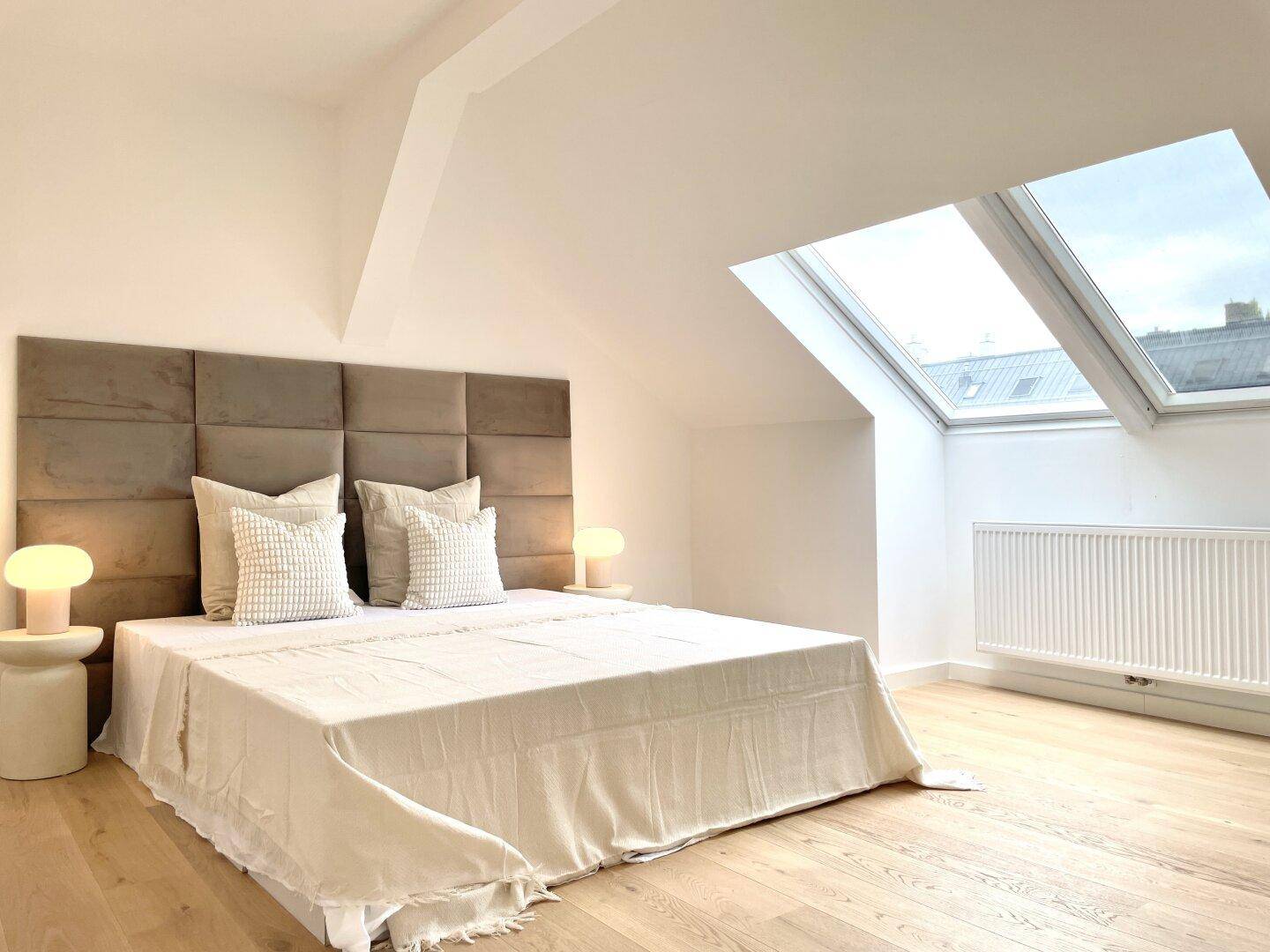 Schlafzimmer mit großem Bett, gepolstertem Kopfteil und Heizkörper unter den Dachfenstern.