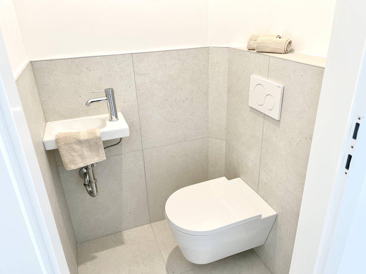 Modernes Gäste-WC mit wandhängender Toilette, kleinem Waschbecken und hellen Fliesen.