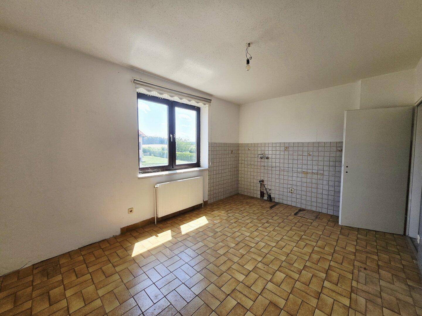 Heller Küchenraum mit Fenster, Fliesenboden und vorbereiteten Anschlüssen für eine Küche.